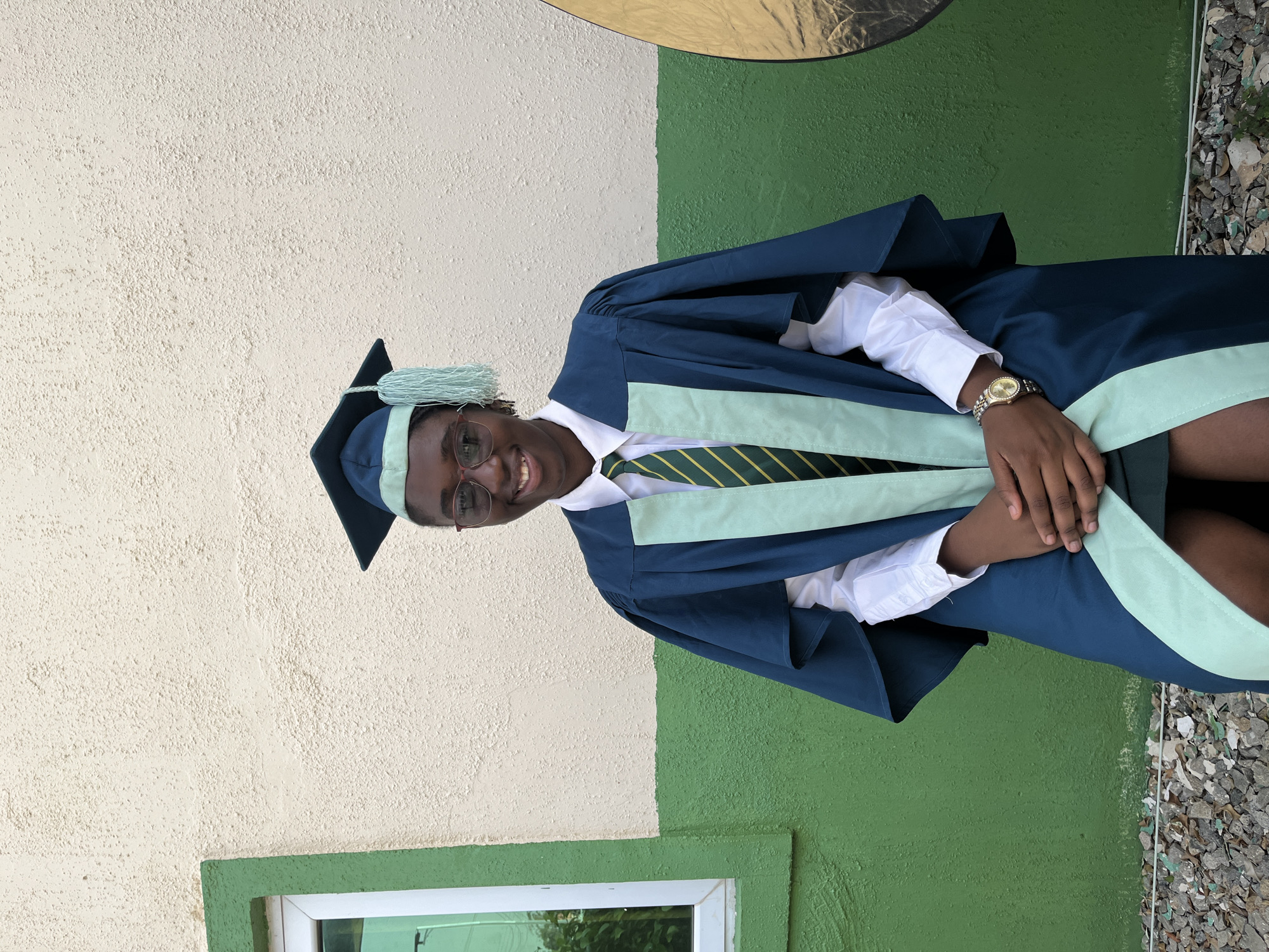 A young man is the subject of this full shot. He is dressed in academic attire—a navy blue graduation gown with a light green trim, a matching graduation cap with a tassel, and a white shirt with a striped tie. He's also wearing glasses and a gold watch. He is smiling, with his hands clasped in front of him.
The background is a wall painted in two tones: the top half is cream, and the bottom half is green. A window with a green frame is visible in the lower-left corner, with plants partially visible through the window. The ground on the right side has loose gravel and bits of debris. A gold reflector is positioned in the upper-middle part of the frame.
While the city of Jos, Nigeria is mentioned, there is no information in the image to verify that this photo was taken there. Therefore, I cannot confirm the location.