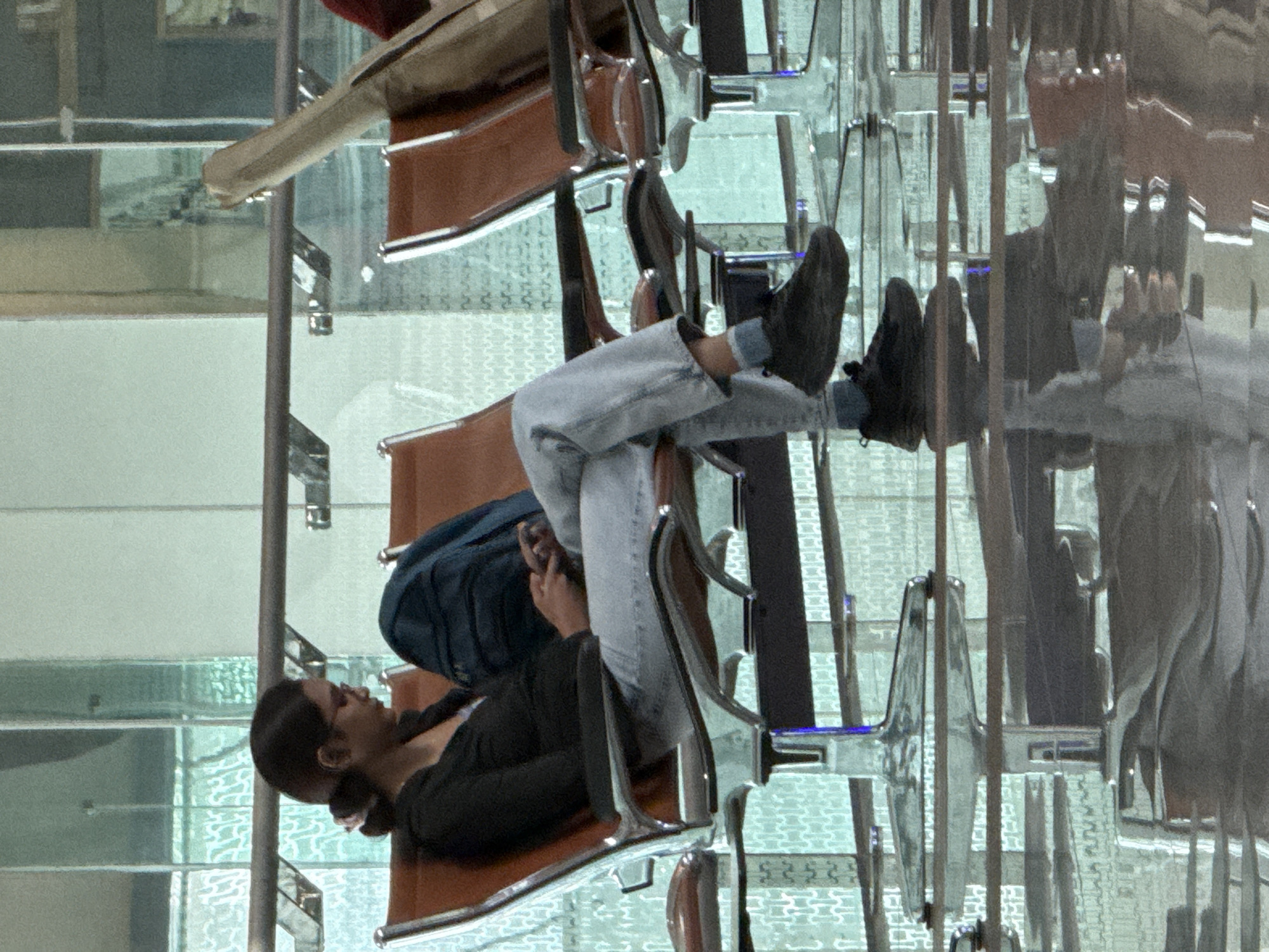 A young woman with dark hair tied back in a bun and wearing glasses is sitting in a row of airport chairs, looking down at her phone. She is wearing a black long-sleeved shirt, light blue jeans, and black sneakers. A dark blue backpack is placed behind her on the chair. The airport waiting area has shiny, reflective floors and a patterned glass wall. The scene appears to be in Seeb Province, Oman, likely at the Muscat International Airport, given the architectural style and the context of a travel hub. The lighting is bright, suggesting it's daytime.
