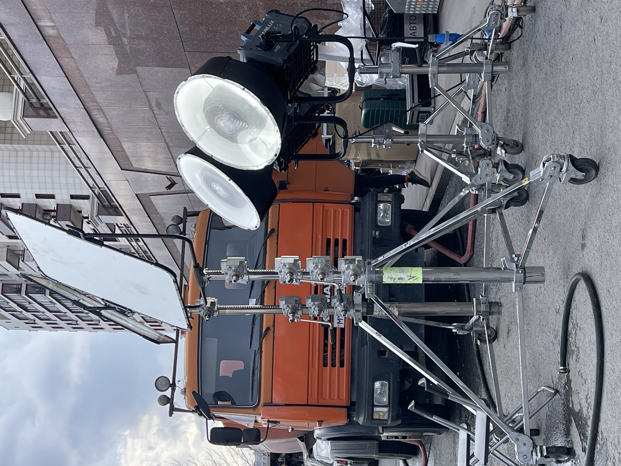 The image depicts a large orange truck parked on a street in what appears to be Moscow, Russia, based on the architectural style of the buildings in the background. The truck is outfitted with extensive lighting equipment, including two large circular lamps, a light-diffusing panel, and complex chrome rigging on wheels. The equipment is for film or television production. The scene appears to be set up for filming outdoors.
