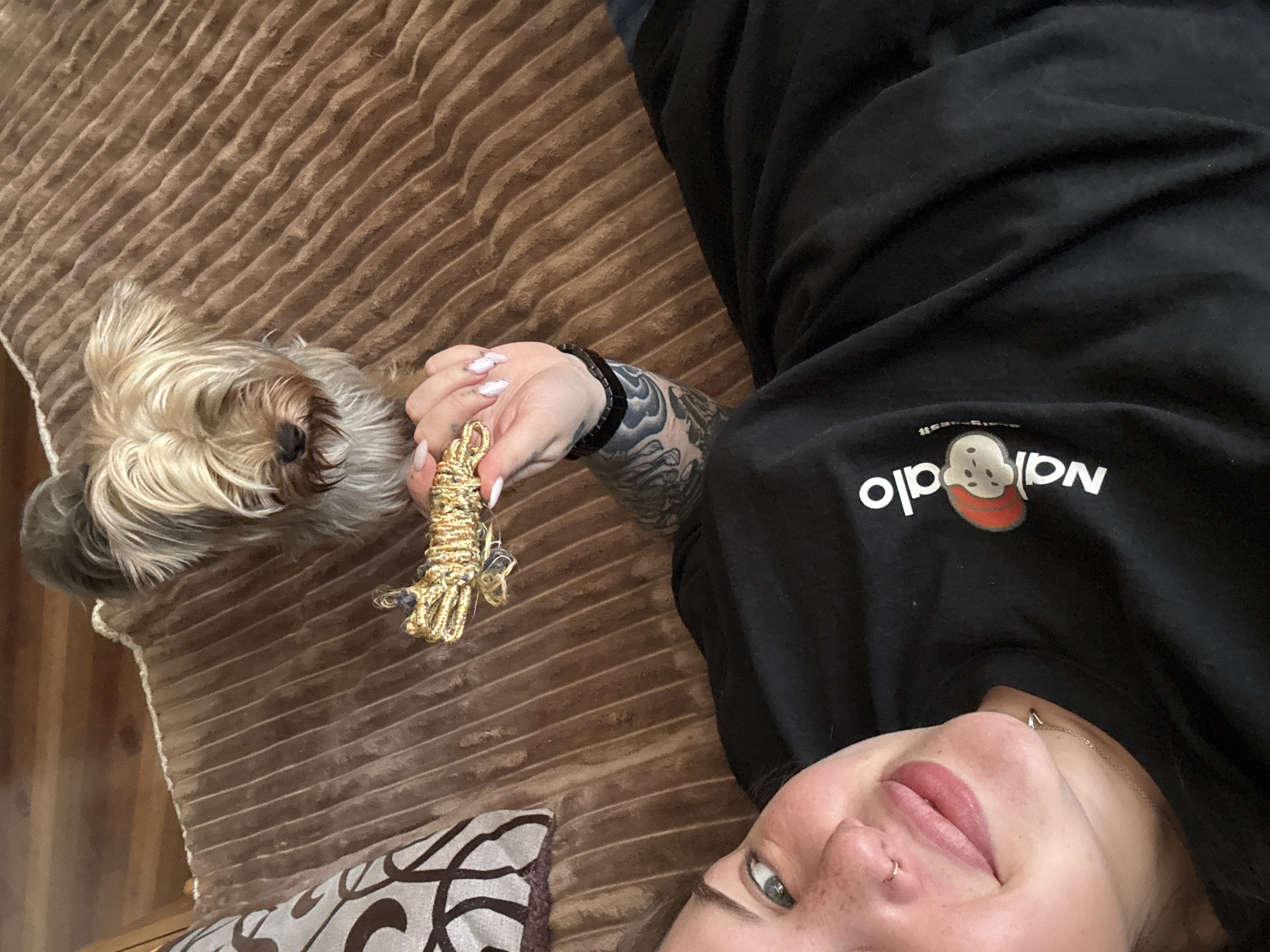 A woman lies on a brown corduroy sofa. A dog is positioned beside her, near her left hand, and she holds a gold-colored, rope-like object in her fingers. The woman has tattoos on her left arm, a nose piercing, and freckles. She is wearing a black sweatshirt with the word "Nala" visible in white print. The location is unknown and it is not clear that it is in the city of Chornomorskoe, Ukraine.