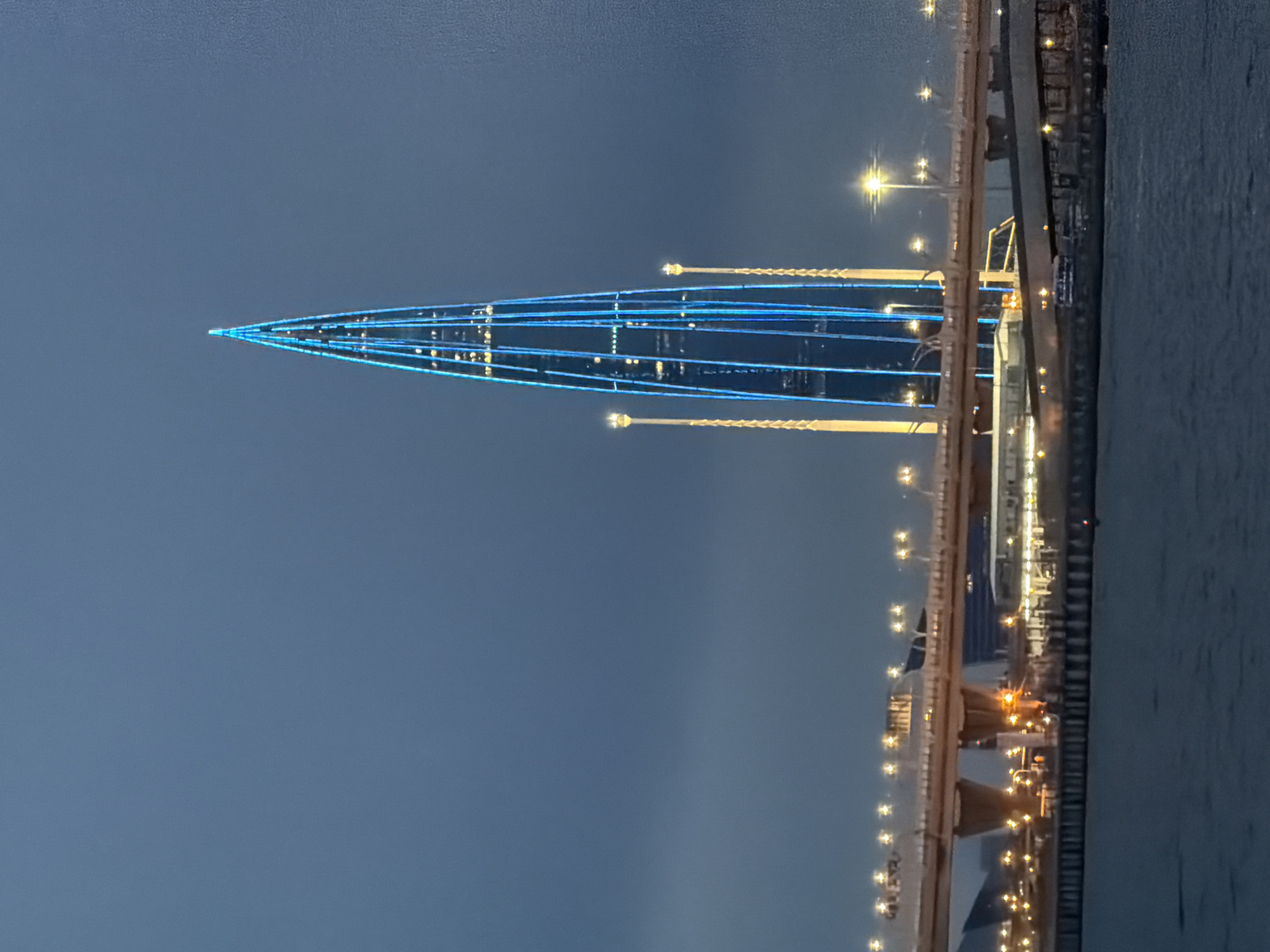 This media file captures a nighttime view of the St. Petersburg stadium, locally known as "Gazprom Arena," illuminated by blue lights. The stadium, with its distinctive pointed, conical shape, is the central subject, appearing to pierce the dusky sky. Strands of bright blue lights outline its structure, creating a striking visual against the dark, hazy atmosphere.
In the foreground and extending into the background is a body of water, likely the Neva River or a bay. Along the right side of the frame, a dock or pier is visible, lined with lights that reflect on the water's surface. Several vessels, possibly ships or ferries, are moored at the dock, their windows and deck lights also glowing. The sky is a deep, muted blue, suggesting it is either very late at night or early morning, with no visible stars or moon. The overall mood is serene and a bit industrial, dominated by the artificial illumination of the stadium and the urban waterfront. There is no visible text in the image.
