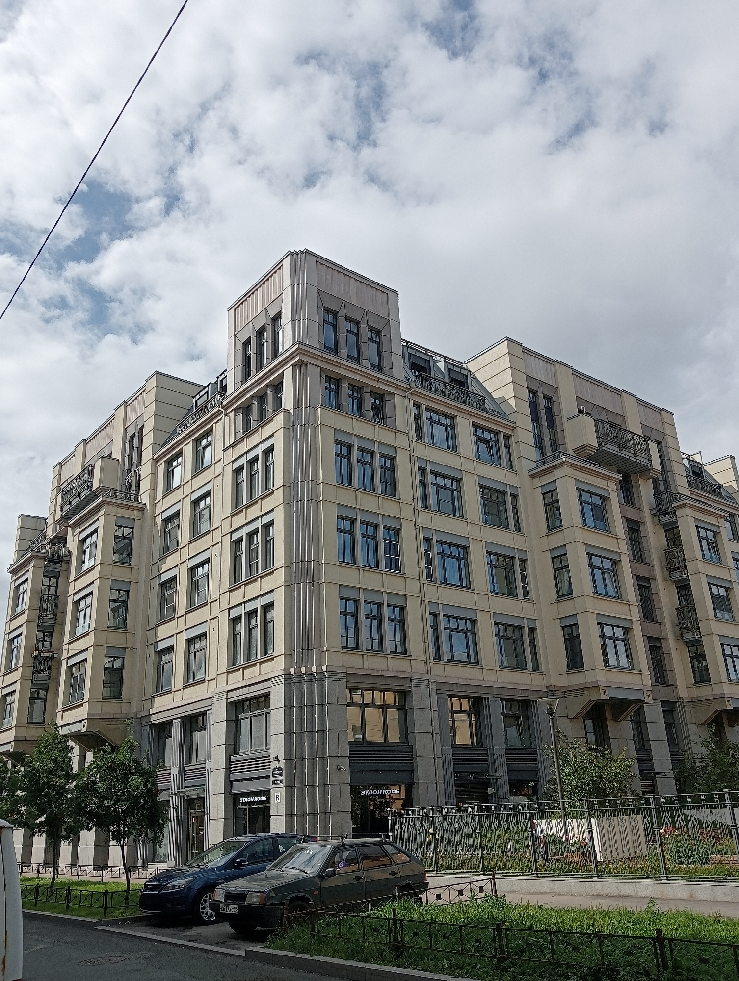 This is an exterior, eye-level, full-shot photograph of a modern, multi-story building located in Saint Petersburg, Russia. The building is constructed with a pale beige facade and features numerous windows with dark frames, some with balconies. The architectural style appears to be contemporary with classical influences, evidenced by the decorative vertical elements on the corners and the cornices. There are several trees in front of the building, and a black metal fence runs along the sidewalk. Two cars, a dark blue sedan and a dark green sedan, are parked on the street in front of the building. The sky is partly cloudy with a few dark clouds. A thin wire runs diagonally across the upper left quadrant of the image. The scene is set on a street, with grass and some vegetation visible along the sidewalk and in front of the fence. The signage on the building indicates a business called "ЭТЛОН КОФЕ" (Etlon Coffee).