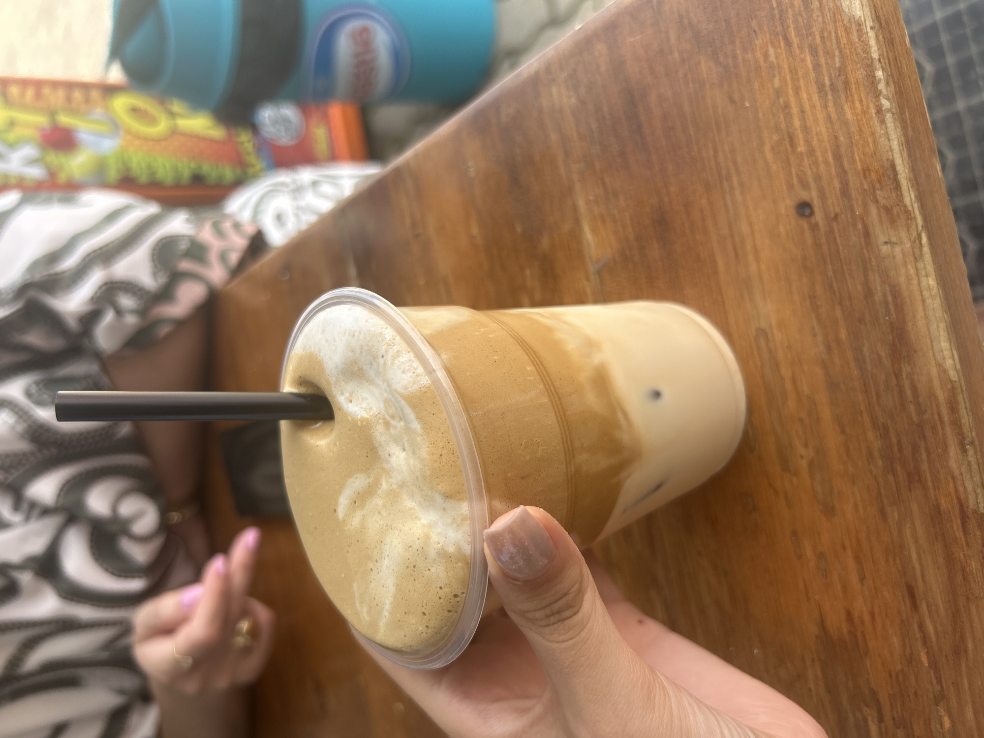 A hand, with light-toned skin and neutral-colored nail polish, holds a clear plastic cup containing a layered beverage. The drink has a thick, foamy, light brown top layer, followed by a darker brown layer, and a light beige liquid at the bottom with visible ice cubes. A dark straw protrudes from the foam. The cup rests on a wooden table with a visible grain. In the background, out of focus, is a person wearing a black and white patterned top. To the right, partially visible, are a bright yellow and orange package and a blue cylindrical object. The scene is likely outdoors or in a casual setting.