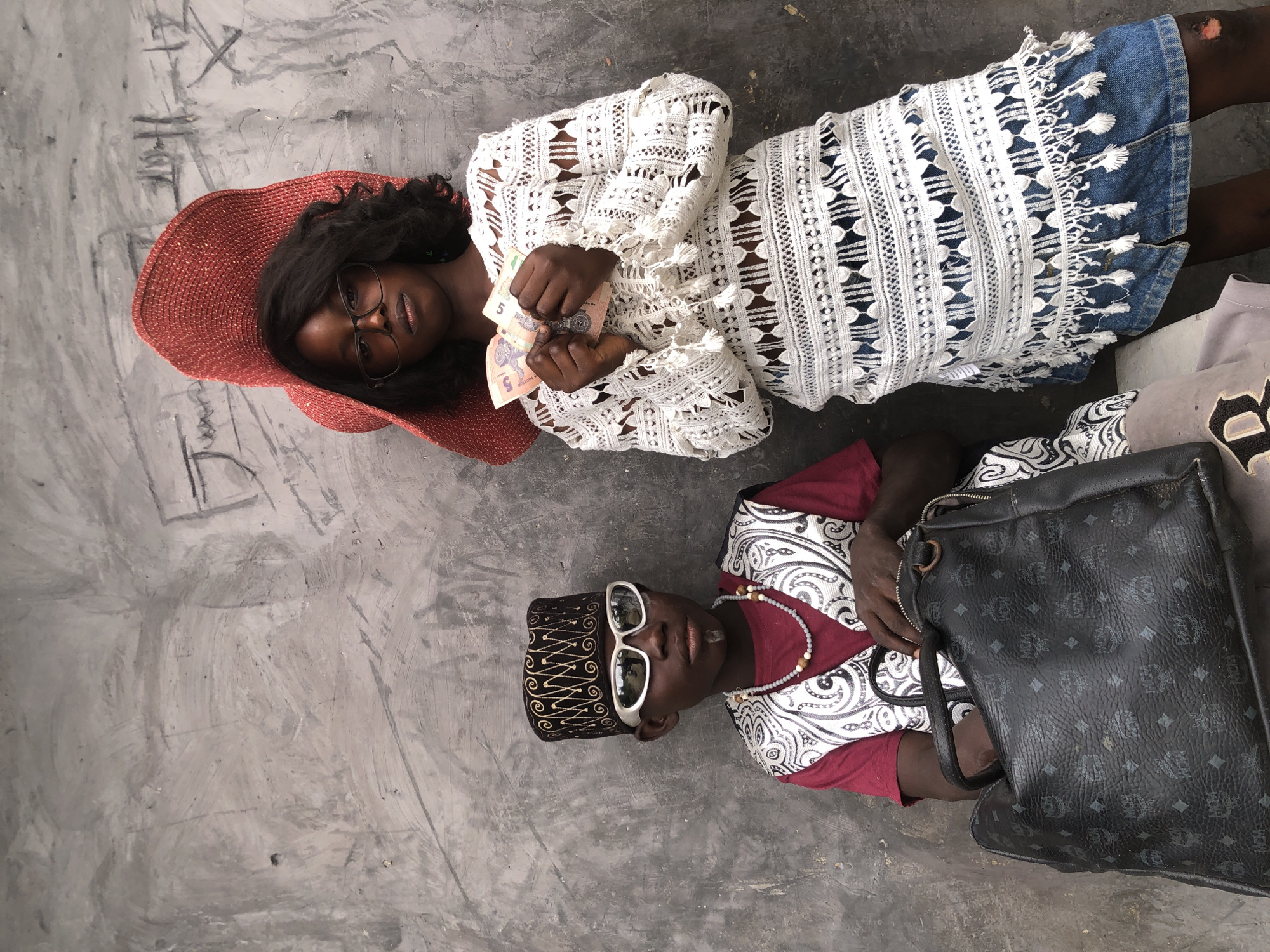 Two children, a boy and a girl, are posing in front of a textured, grey wall in Ado, Nigeria. The boy, on the left, is wearing a traditional cap with gold embroidery, large white sunglasses, a maroon t-shirt, and a patterned vest. He also has a goatee and is wearing a beaded necklace. He is sitting next to a large, black designer bag with the MCM logo.
The girl, on the right, is wearing a wide-brimmed red hat, glasses, and a white lacy top with fringe detailing over a denim skirt. She is holding several banknotes in her hands. The wall behind them has some faint markings and graffiti on it. The lighting suggests it is daytime.