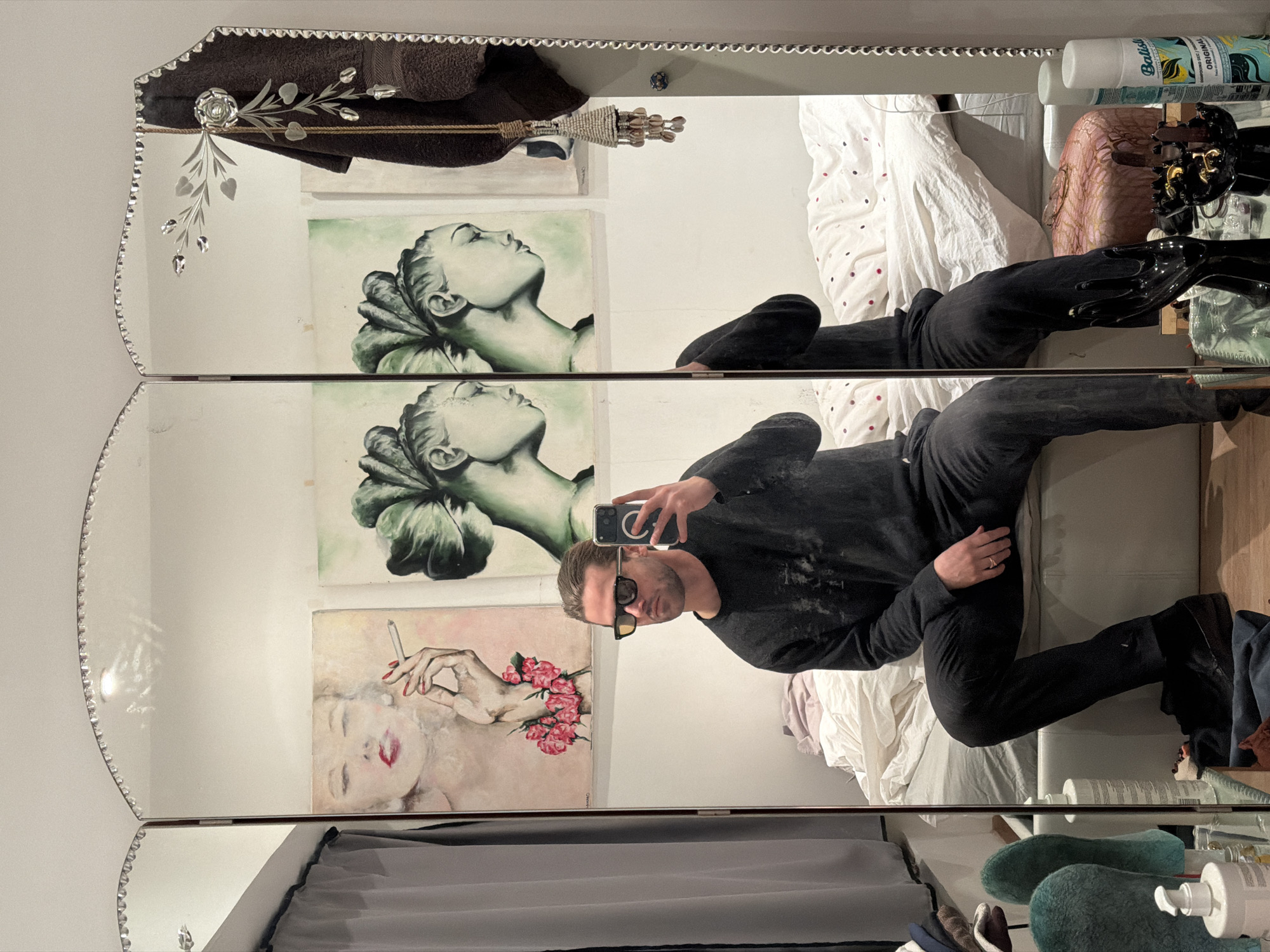 The scene depicts a male subject taking a selfie in a decorative tri-fold mirror within a private room in Paris, France. The man, with light brown hair pulled back, wears dark sunglasses with amber-tinted lenses, a dark long-sleeved shirt, and dark trousers. He is seated, holding a smartphone (identified by its visible camera and MagSafe ring) in his right hand, capturing his reflection. A metallic ring is present on his left ring finger.
The mirror features a scalloped top edge adorned with small clear beads and an etched floral design on its rightmost panel. Reflected in the mirror, behind the man, are three framed artworks mounted on a white wall: on the left, a painting of a woman with red lips holding a cigarette, with pink roses around her arm; in the center and right, two identical profile paintings of a woman with an elaborate green coiffure against a light green background.
Below the artworks, a bed with white bedding patterned with small red and dark polka dots is visible. To the far left, a grey curtain is partially drawn. On a surface below the mirror, various objects are present: two teal-colored, textured, decorative forms, a white pump bottle, a black jewelry display shaped like a hand, a smaller black decorative dish, and two aerosol cans, one identified as "Batiste Original Dry Shampoo." A dark brown towel hangs draped over the upper right side of the mirror frame, from which a beaded tassel with shell-like elements also hangs. The environment is consistent with a bedroom or dressing area.