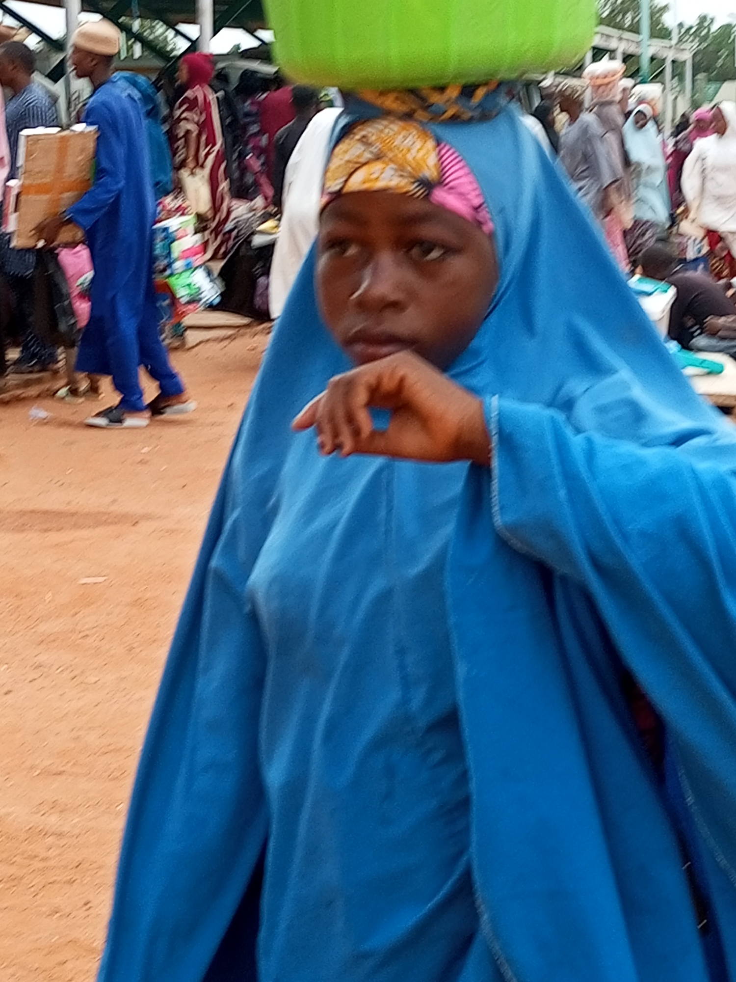 In a bustling marketplace in Gombe, Nigeria, a young girl with dark skin and a vibrant blue hijab and dress is the central focus of the image. She is carrying a large, light green bowl balanced precariously on her head, secured by a colorful patterned wrap. Her face is turned slightly away from the camera, her gaze directed downwards, giving her a pensive expression. Her left hand is raised, resting on her chest, with her fingers delicately curved, as if in thought or speech.
The background reveals the lively atmosphere of the market. To her left, a man in a blue robe and light-colored cap carries a cardboard box. Other figures in the background are partially obscured, suggesting a dense crowd of shoppers and vendors. The ground is covered in reddish-brown earth, typical of the region. Behind the people, stalls are visible, displaying a variety of goods, including textiles and what appear to be packaged items. The overall scene is one of everyday life unfolding in a vibrant, public space.