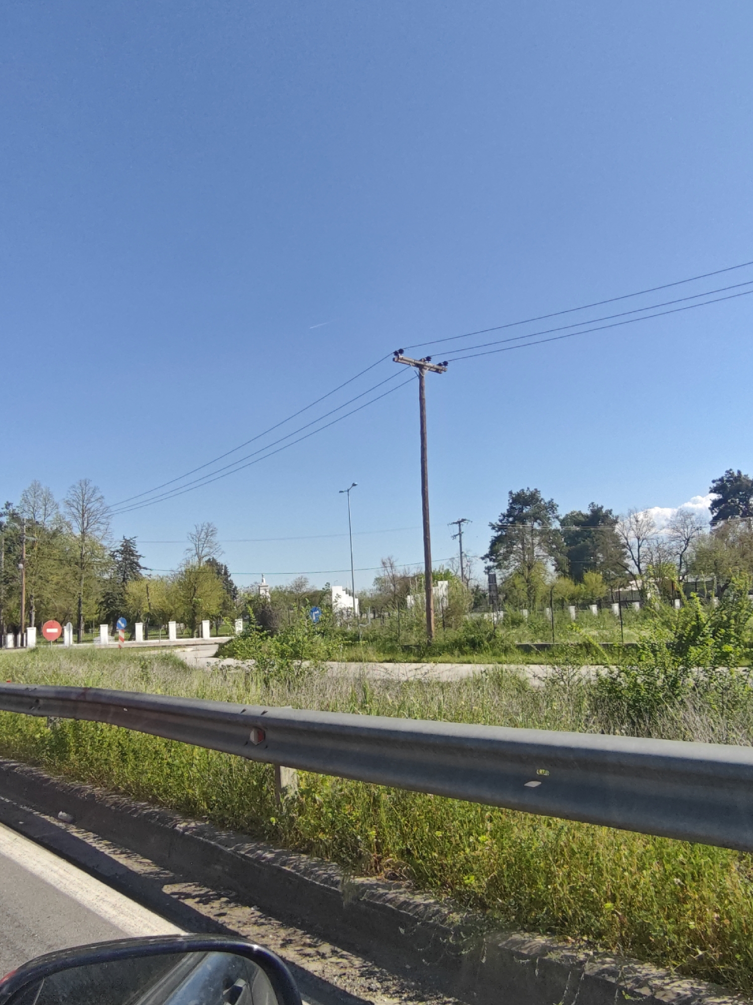 The image depicts a view from a moving vehicle, likely on a road in Serres, Greece. A gray metal guardrail is visible in the foreground, with vegetation growing alongside it. Beyond the guardrail, there is a grassy area with scattered bushes and trees. In the background, a white building with a spire can be seen, likely a church or religious structure. Utility poles and power lines are visible against a clear blue sky. Road signs are positioned along the roadside, some with visible red circles. The image is taken during daylight hours.