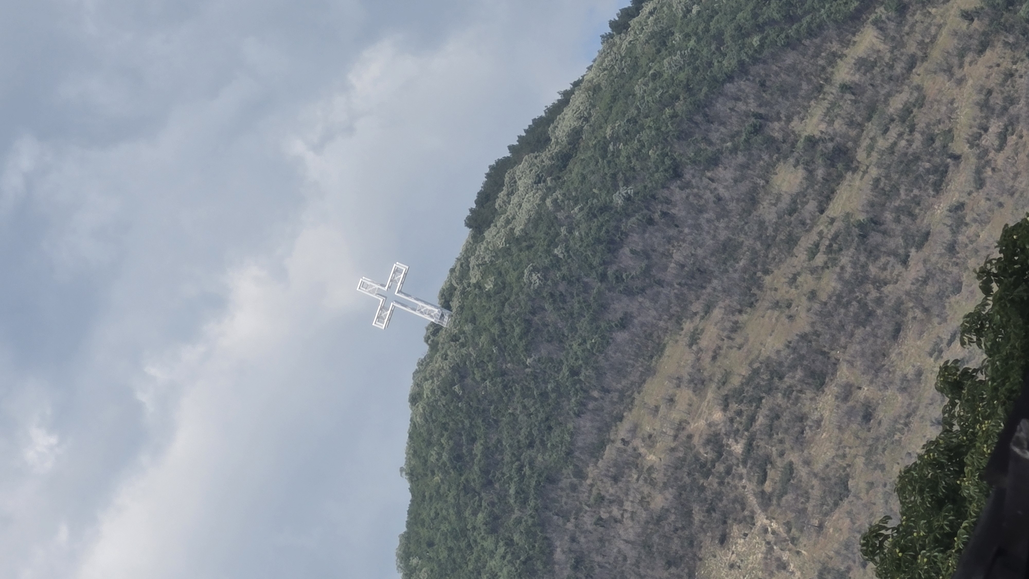 A large, white, illuminated cross is positioned at the summit of a tree-covered mountain. The cross is centrally located in the frame, standing out against a cloudy, overcast sky. The mountain slopes downwards, with a mixture of dense green vegetation and sparser, dry brush. The location is the Karandila mountain in Sliven, Bulgaria.