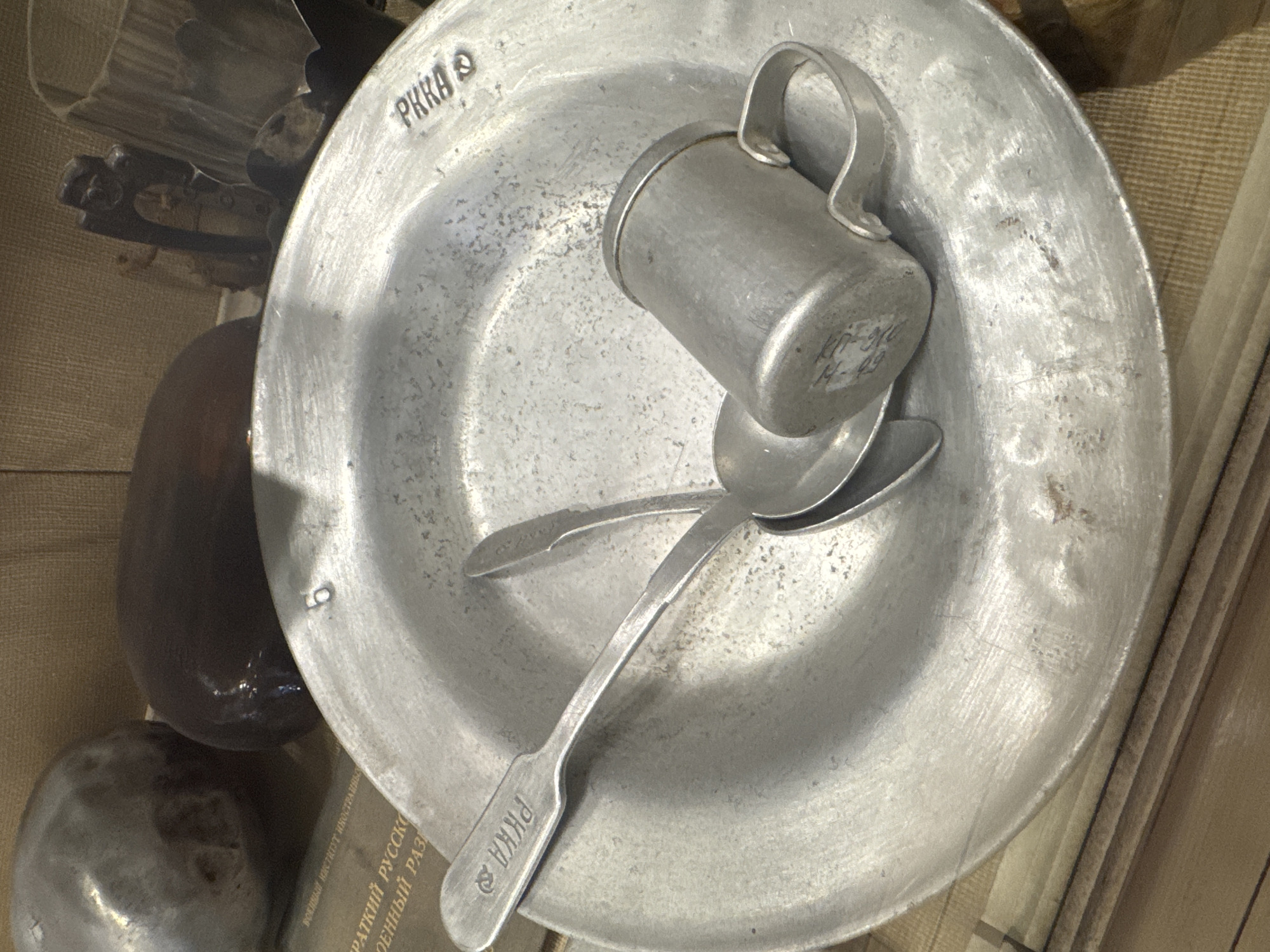 The image shows a collection of metallic eating utensils, likely an exhibit in a museum. A shallow, round plate made of a dull silver metal dominates the scene. The letters "PKKA" are stamped near the top edge of the plate, and again on the handle of a spoon that rests within the plate. A small, cylindrical cup with a handle sits on top of two spoons inside the plate. The cup also appears to be made of the same metal as the plate, and has some markings on its side. The scene is lit by a soft, diffused light, and other indiscernible objects are partially visible on the left side of the frame. The bottom of the frame shows part of text with the beginning "ВОЕННЫЙ ИНСТИТУТ РУССКОЙ", suggesting a military context. The scene is likely inside a museum or display case. Given the Cyrillic text, the location is likely Saint Petersburg, Russia.