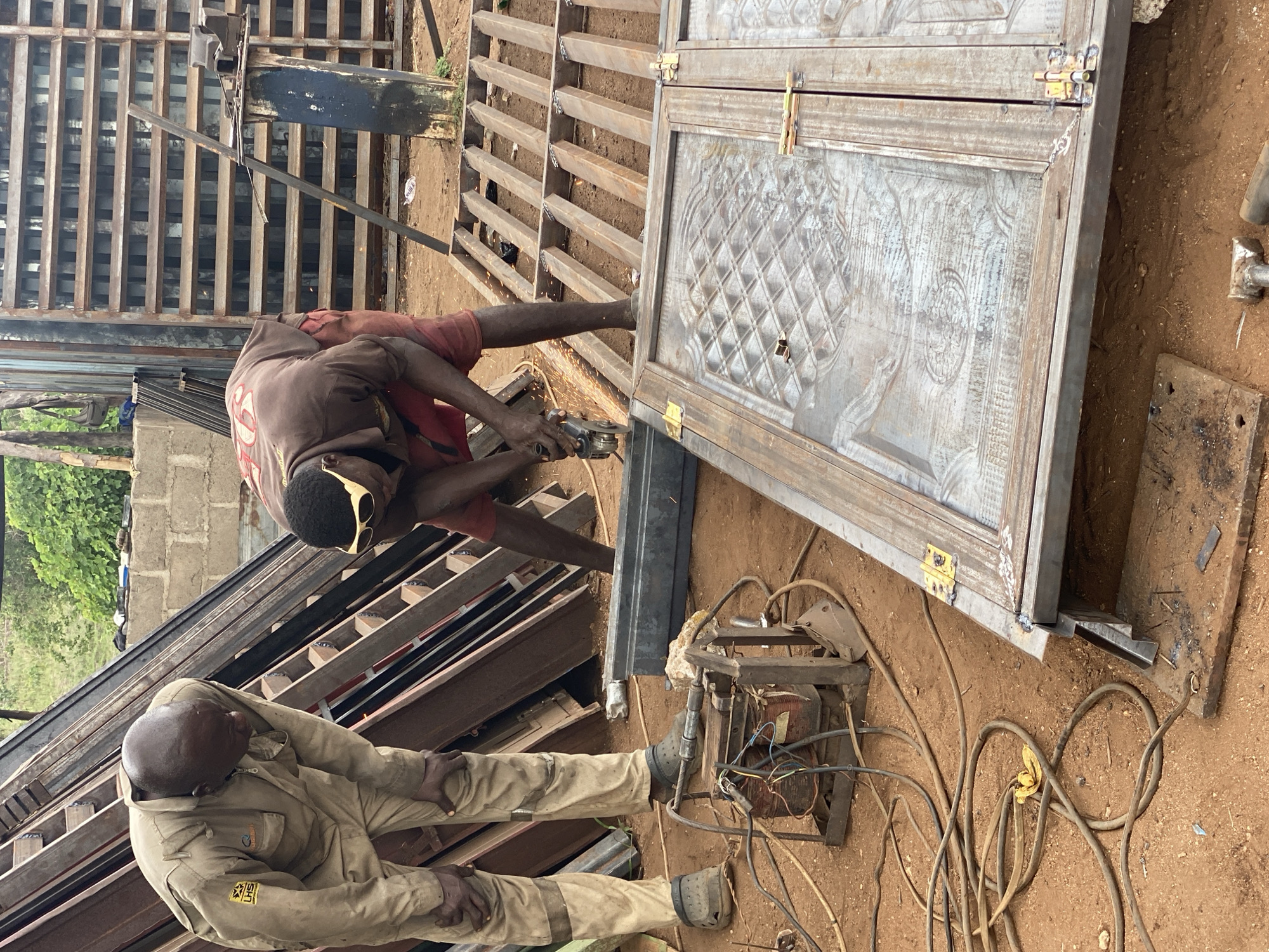 The image depicts an outdoor scene in Adankolo, Nigeria, showing two men working on metal structures. One man, wearing a brown shirt and red shorts with safety glasses, is using a power tool, likely a grinder, on a metal frame. Sparks are visible. The other man, wearing a tan uniform, is sitting on a pile of metal bars. A welding machine with wires is on the ground nearby. A large metal door frame with a patterned interior is also present. The ground is dirt. Additional metal bars and structures are in the background.
