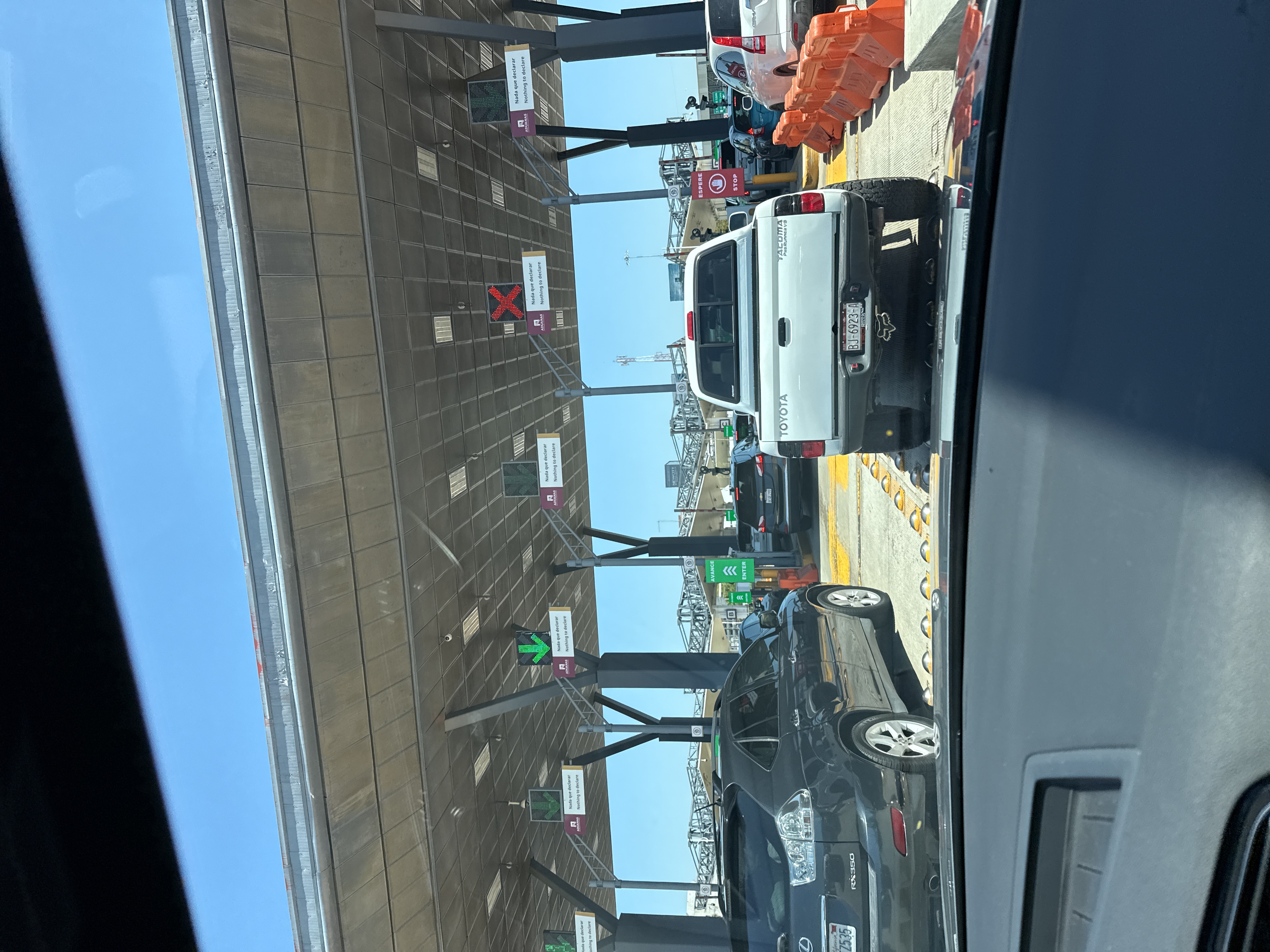 The image shows a queue of vehicles at a border crossing in Tijuana, Mexico. Overhead structures feature signs with green arrows and red Xs, indicating lane availability, along with text that reads "Nada que declarar" in Spanish and "Nothing to declare" in English. Vehicles visible include a dark SUV, a white Toyota Tacoma truck, and a white SUV. The license plate of the Toyota Tacoma is visible. Orange traffic barriers are on the right side of the image. The sky is clear and blue.