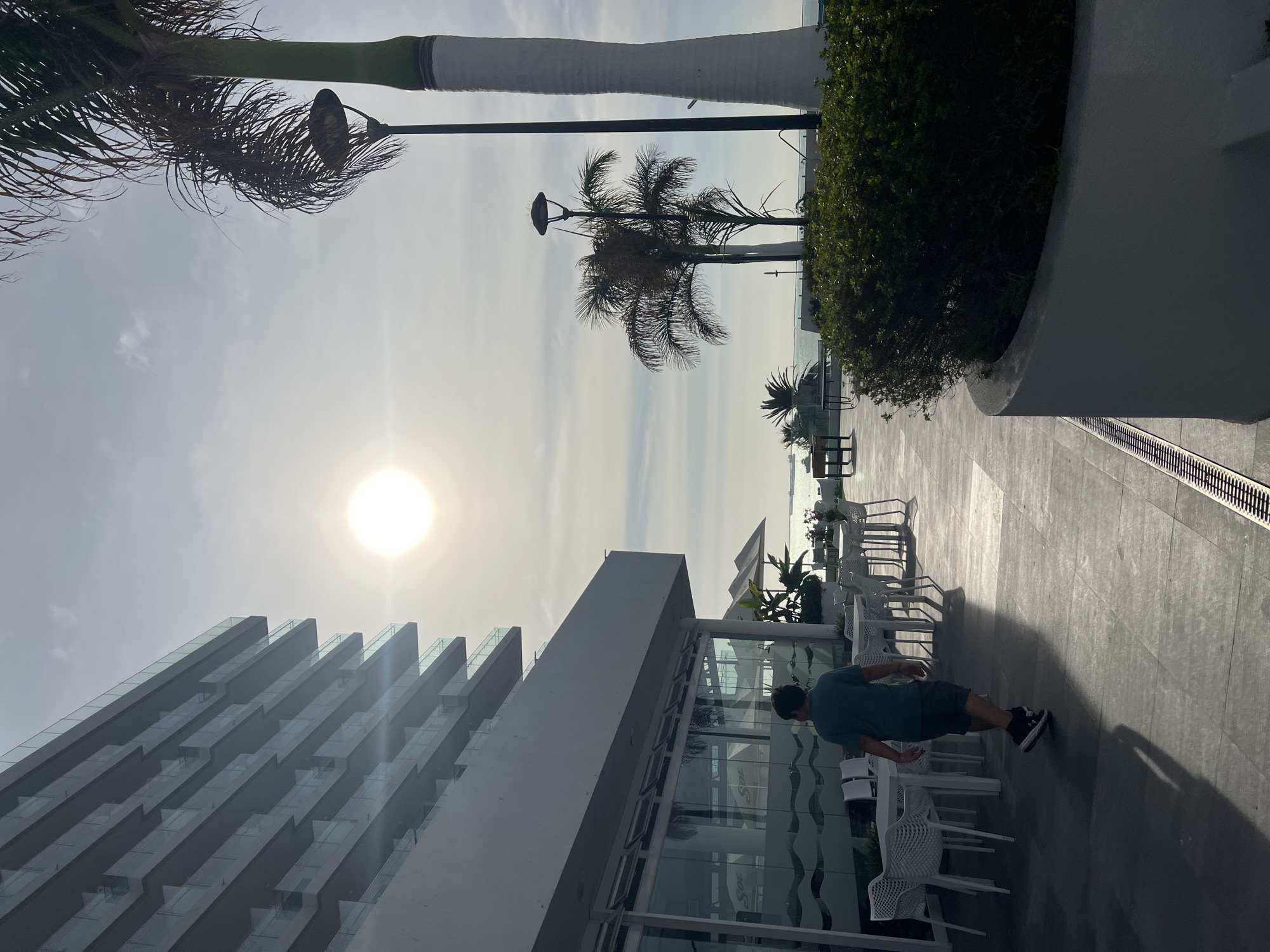A partially obscured wide-angle view, taken outdoors during daylight, shows a person walking on a patio or walkway adjacent to a modern building. The location is strongly suggested to be the city of Arica, Chile. The sun is visible and high in the sky, causing lens flare.
**Objects:**
* A modern building with reflective glass and angular architecture.
* A walkway or patio paved with gray stone or concrete.
* White plastic patio chairs and tables.
* Tropical foliage including palm trees and bushes.
* Light posts.
* A metal drainage grate built into the patio surface.
* A body of water, likely the ocean, is visible in the distance.
**People:**
* A single person is walking on the patio. They are wearing a blue shirt, dark shorts, and black shoes. They are casting a shadow.
**Actions/Interactions:**
* The person is walking in a general rightward direction.
**Environment:**
* The setting is a sunny day, likely a resort or tourist area.
* Based on the architectural style and tropical vegetation, it can be located in Arica, Chile.