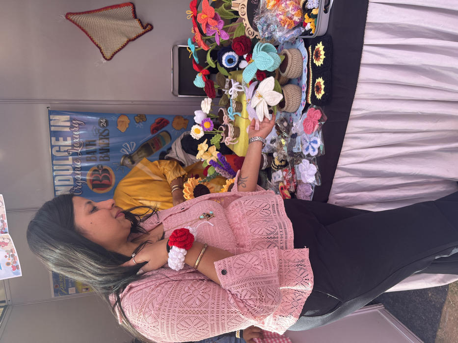 The image shows a woman in a pink lace top and black pants standing in front of a stall selling handmade crocheted flowers. The stall is located in Sector 17, Chandigarh, India (latitude: 30.764, longitude: 76.798). The woman is looking at the crocheted flowers, and one of her hands is reaching out to touch a crocheted flower in a brown pot. The flowers are arranged in a variety of colors, including pink, red, blue, white, and yellow. The woman is wearing a bracelet on her left wrist and a necklace. The stall has a white tablecloth and a black table top. In the background, there is a sign that says "Indulge in Organic Luxury Bath Bakers 24". There is a person in the background wearing a yellow shirt. The stall is located in a market setting, with other stalls visible in the background.