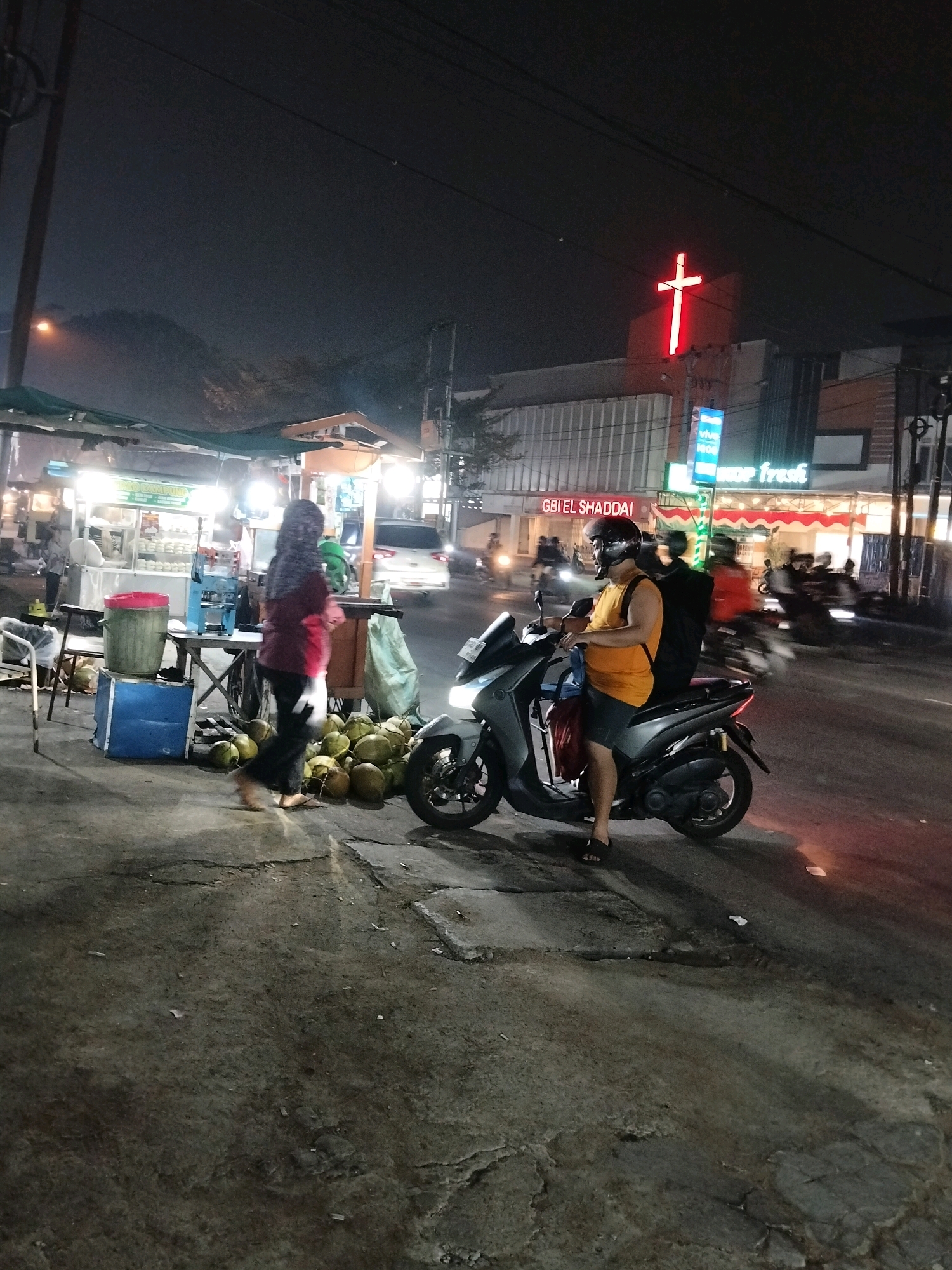 The image captures a street scene at night in Pontianak, Indonesia. A vendor stall is visible on the left, with various food items or goods displayed under a green awning. A person in a dark headscarf and maroon top walks past the stall, carrying a white bag. In front of the stall, a pile of coconuts is present, along with a large green bin and a blue crate. A person wearing a yellow shirt and a helmet is seated on a scooter in the foreground, facing towards the right side of the image. Several other scooters and motorcycles are on the road in the background, some with riders. A prominent red neon cross illuminates the facade of a building on the right side of the frame, identified by signage as "GBI EL SHADDAI" and "VIVE FRESH". The overall environment suggests a bustling urban area with food vendors and traffic at night.