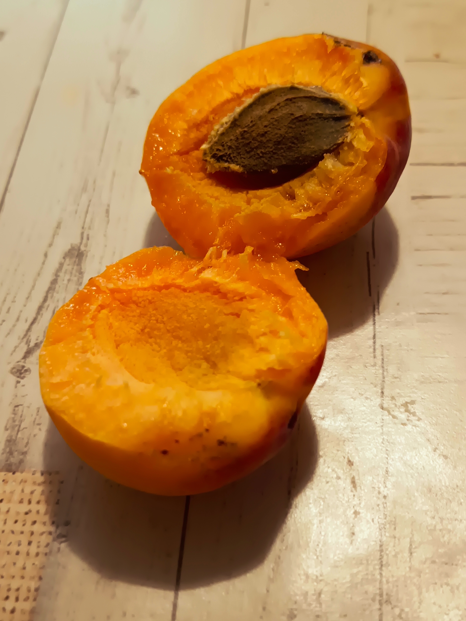 Two halves of a cut apricot are placed on a wooden surface. The apricot on top is cut in half, revealing its seed, which is brown and textured. The flesh of the apricot is a vibrant orange with some red blush on the skin. The apricot on the bottom is also cut in half, showing its juicy, textured interior. The wooden surface has a light color with visible grain patterns and some darker lines. A shadow is cast from the apricots onto the surface. The overall scene is a close-up, still life shot. There is no indication of a specific location in Izmail, Ukraine.