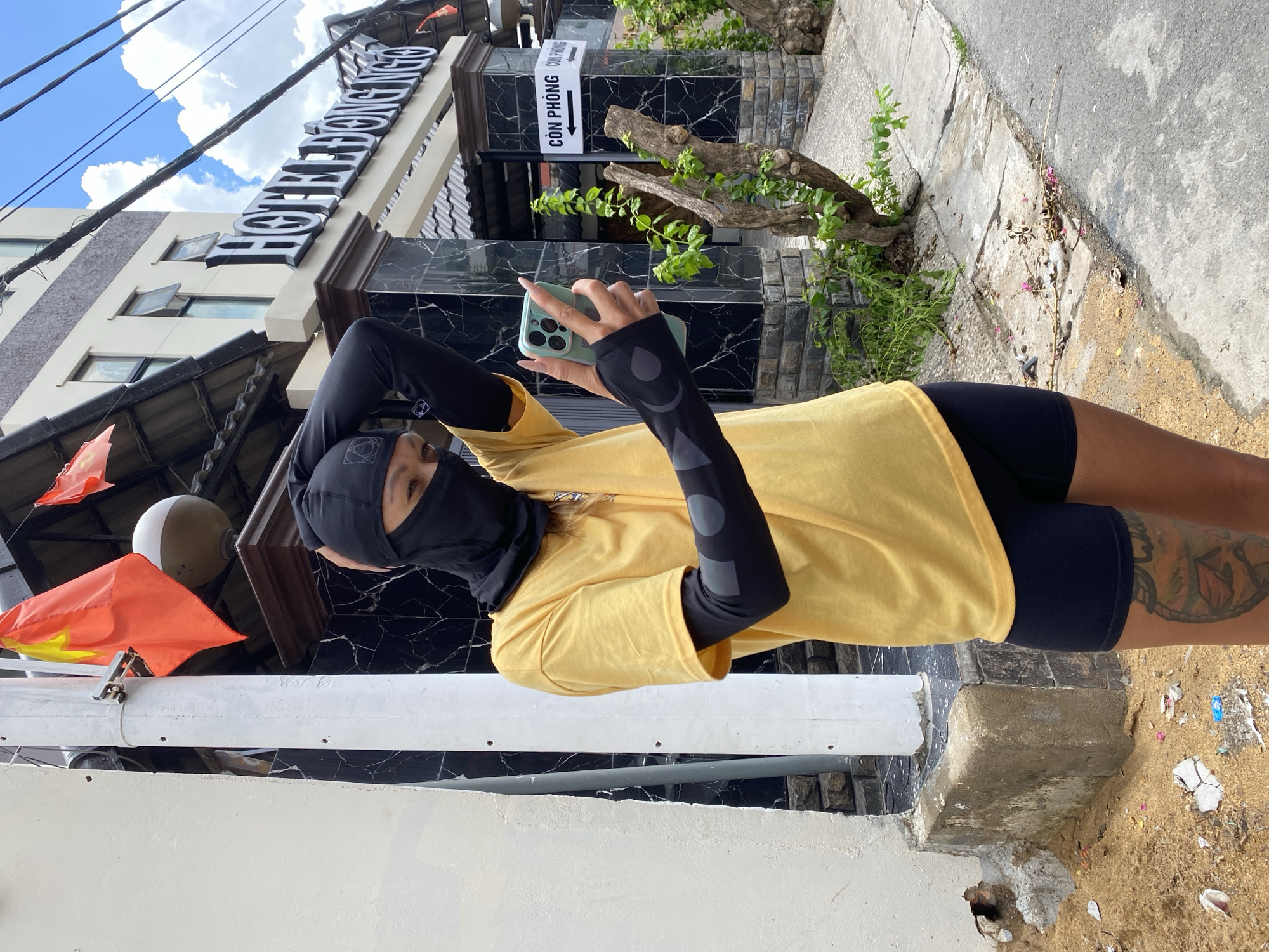 A person wearing a yellow t-shirt, black cycling shorts, and a black balaclava is taking a photo with a smartphone. The person is standing on a sidewalk, in front of a building named "HOTEL DONG NAI," as seen on a sign in the background. A sign reads "CON PHONG." A Vietnamese flag is displayed on a pole to the left. The sidewalk is cracked and littered. A small, leafless tree branch is visible against the building.