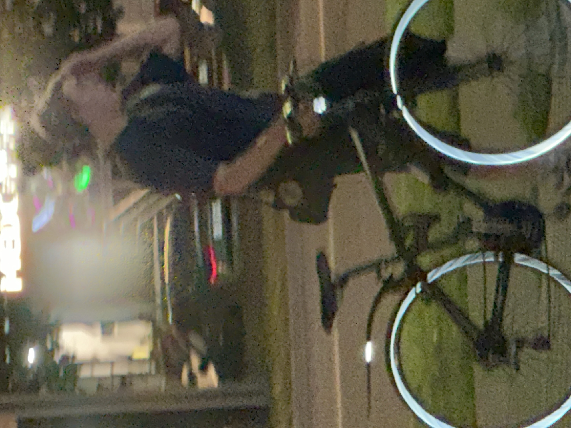 The image depicts a blurry, low-light scene at night on a street, possibly in Podolsk, Russia, given the reference. A person is seen from the waist down, pushing a bicycle. The person is wearing dark clothing and has their hand raised to their head. The bicycle is black and has large, reflective rims on its wheels. In the background, there are indistinct lights and vehicles, including a red taillight of a car. The overall impression is that of a candid, possibly surveillance, photograph due to the quality and focus.