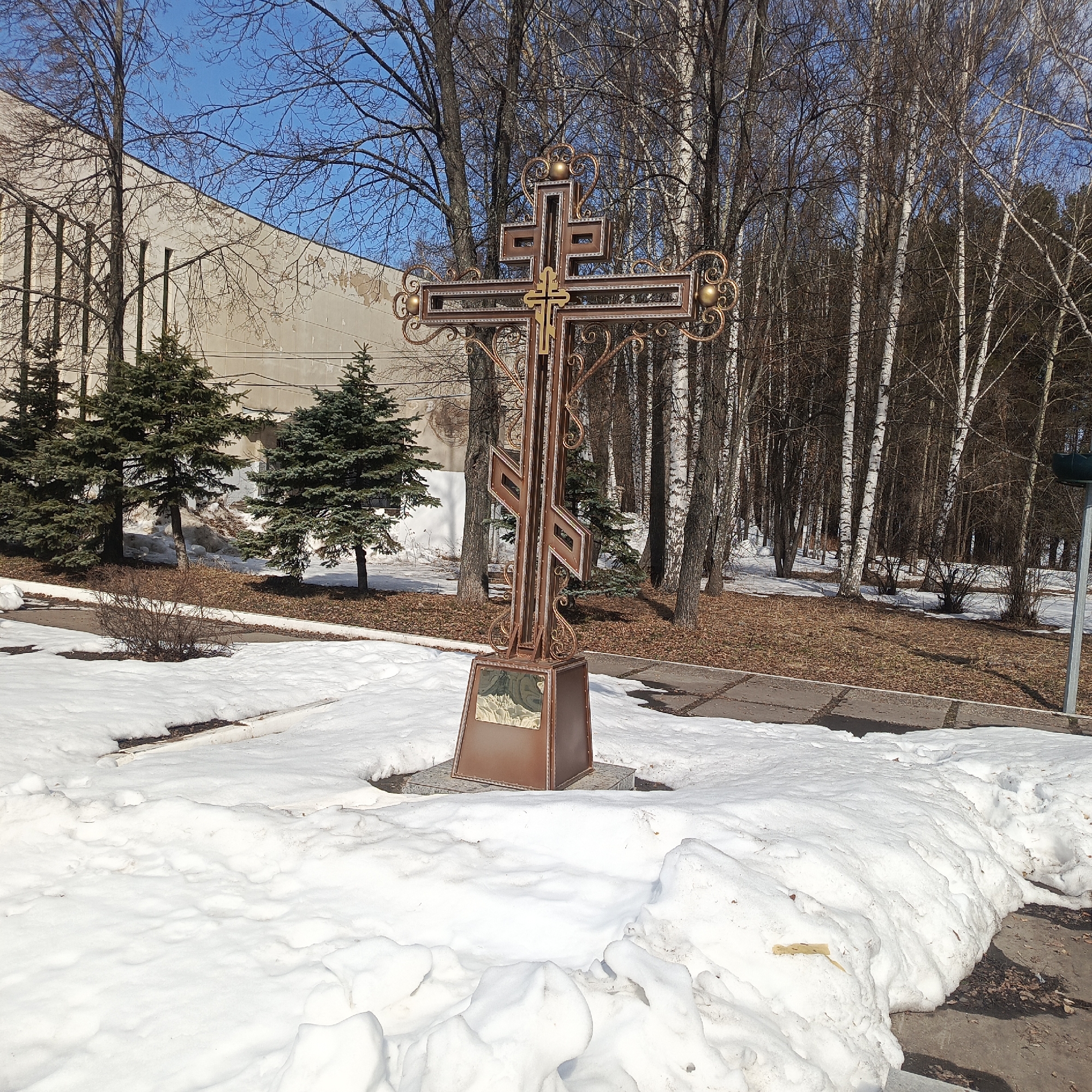 An outdoor scene features a large, ornate Orthodox Christian cross situated in a snowy environment. The cross is made of a dark, bronzed metal with golden accents and is positioned on a pedestal. The ground is covered with snow, with patches of exposed ground and leaves visible. In the background are several trees, including birch trees recognizable by their white bark, suggesting a park or woodland setting. A building with a beige facade is partially visible to the left. The presence of snow indicates winter or early spring conditions.
The cross is located in the city of Chaikovsky, Russia.