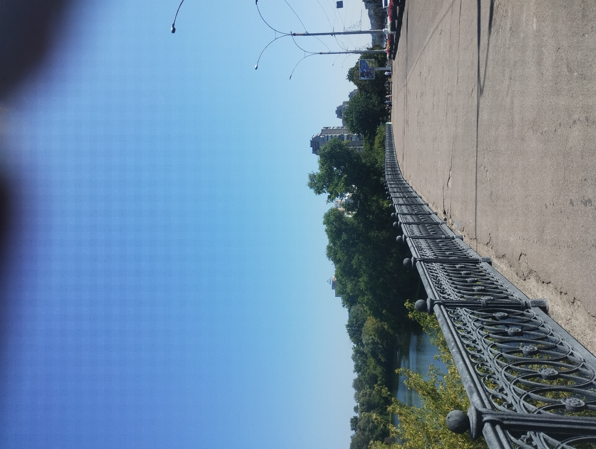 The image shows a view of a pedestrian walkway on a bridge in Sumy, Ukraine. A decorative wrought-iron fence lines the left side of the walkway, separating it from a body of water and lush green trees. In the distance, several multi-story buildings are visible, as well as streetlights and a billboard. The walkway itself is paved with concrete and shows signs of wear, with cracks and patches visible. Several people are walking on the walkway in the distance. The sky is clear and blue, indicating a sunny day.