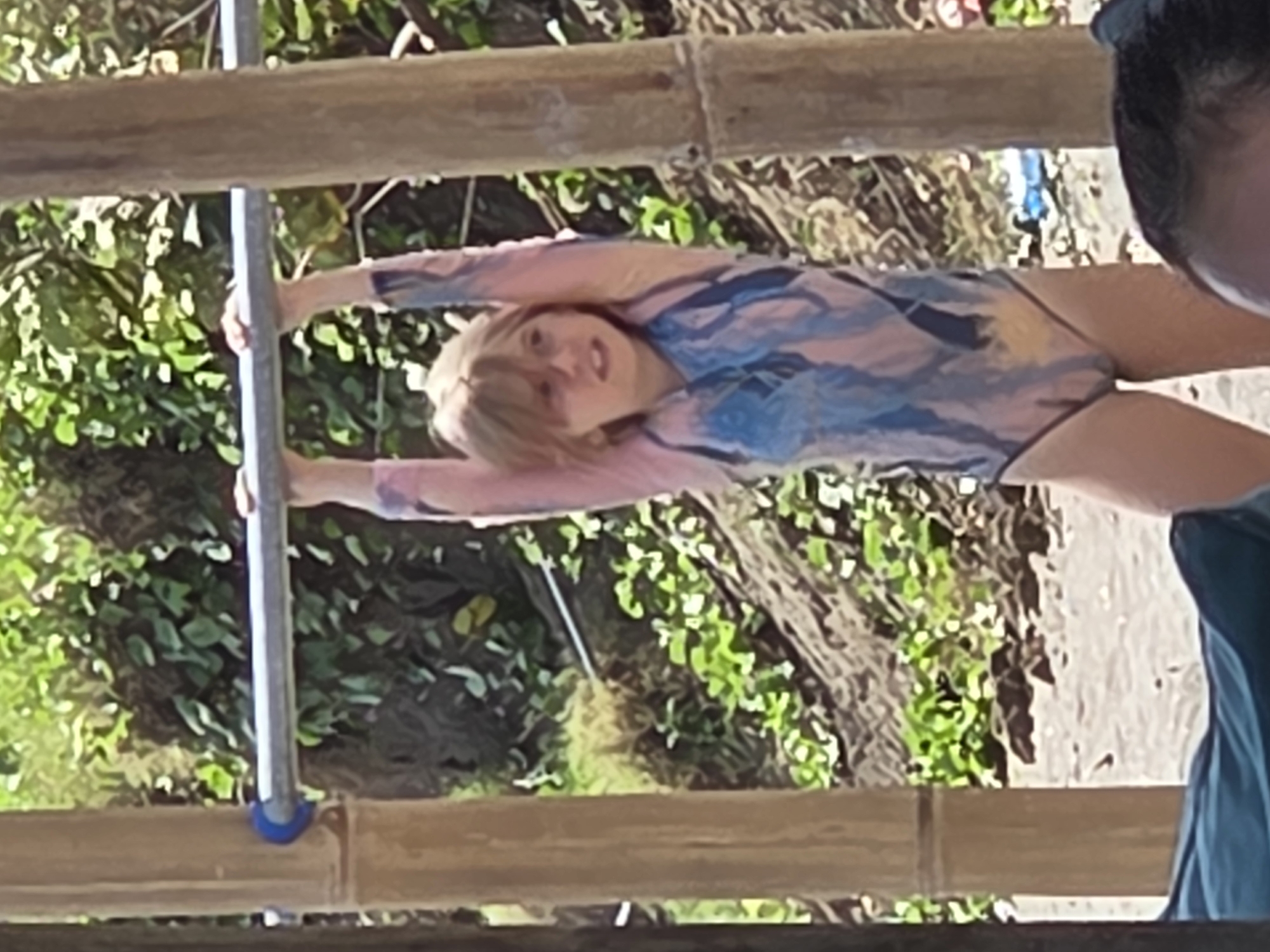 A person with blond hair, wearing a pink and blue long-sleeved swimsuit, hangs from a gray metal pole supported by bamboo beams. The location appears to be outdoors, possibly in North Kuta, Indonesia, based on the tropical vegetation visible in the background. Another person wearing a teal shirt is seen to the right of the frame.