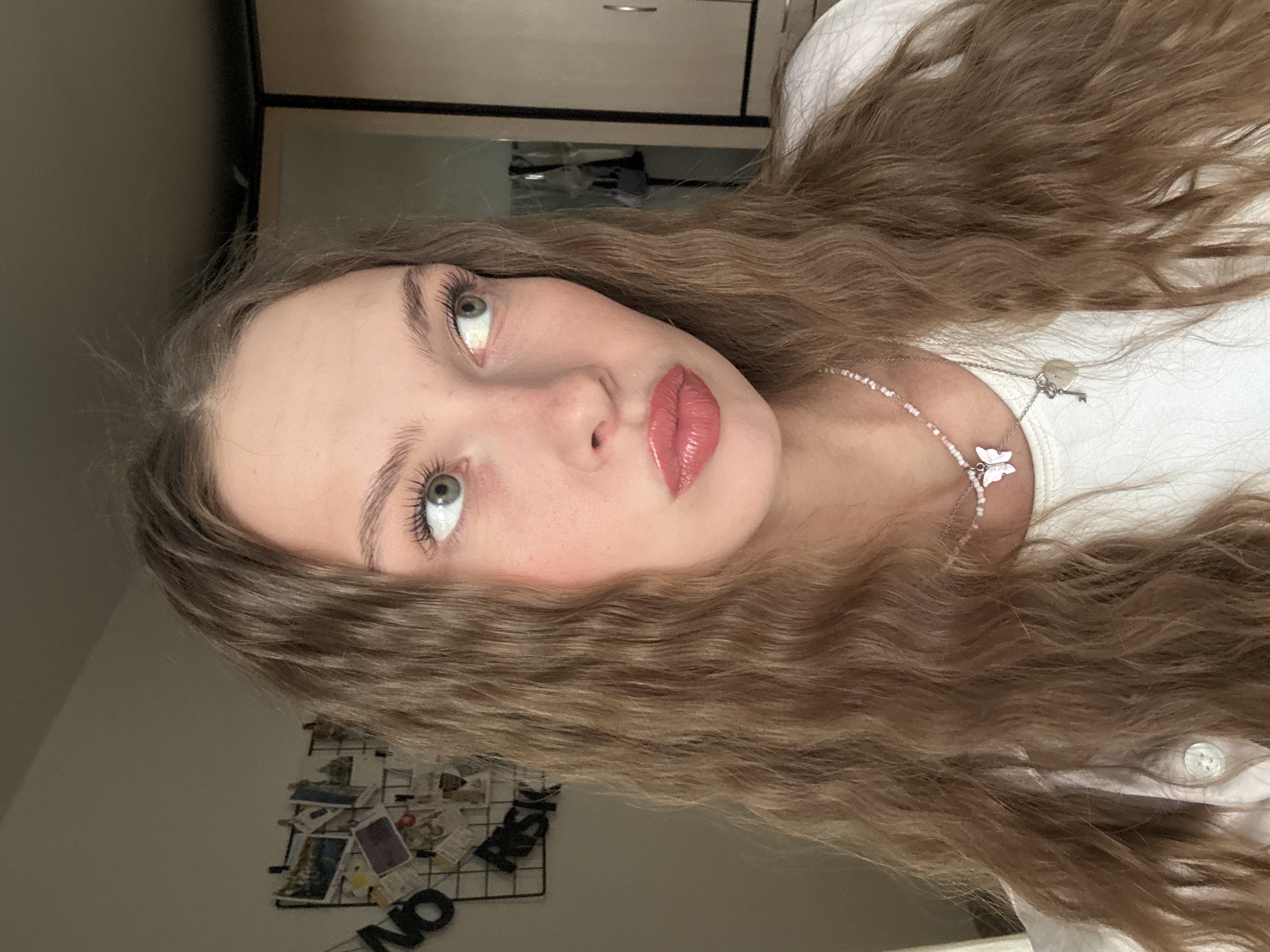 A young Caucasian woman with wavy brown hair and light green eyes is captured in a medium close-up shot indoors. She is looking to her right, with her lips slightly parted in a pout. She is wearing a white t-shirt and a necklace with a butterfly pendant and a key charm. The background includes a wall with a grid of photos and a closet. The lighting suggests an indoor setting, possibly a bedroom, with natural light coming from the right. The image does not provide any information about the city of Perm, Russia.