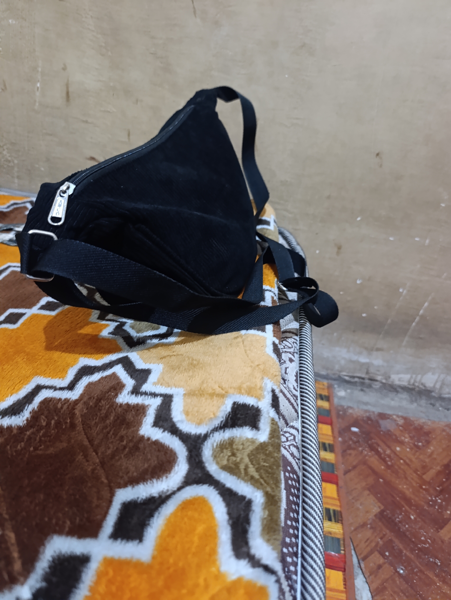 A black corduroy sling bag is placed on a patterned blanket. The bag has a zipper closure and a single strap. The blanket has a geometric pattern in shades of yellow, brown, and white. The background consists of a textured, light-colored wall and a wooden floor. The scene appears to be indoors, possibly in a residential setting. The image does not provide any specific location details for Madatte, Indonesia.