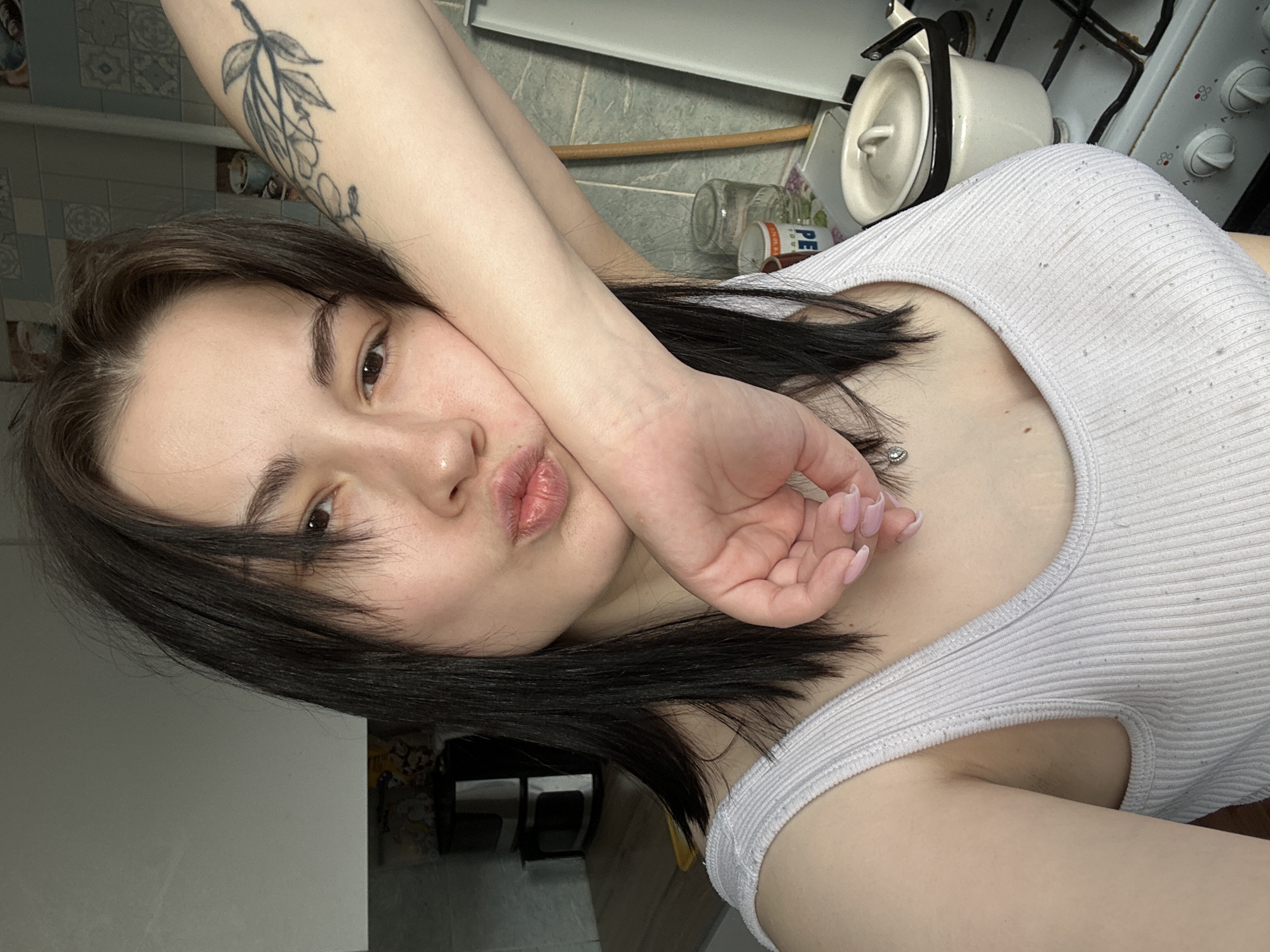 A woman with dark hair, pale skin, and light pink painted nails is in an indoor location, likely a kitchen. She is wearing a white ribbed tank top and has a tattoo on her upper left arm. The woman is posing for the camera with her left hand against her cheek and her lips puckered. Behind her is a kitchen stove with a white kettle on it and various items such as jars and containers. The location is likely a residential kitchen, possibly in an apartment. There is no explicit indication the photo was taken in Chaikovsky, Russia, and no identifying Russian text is visible in the image.