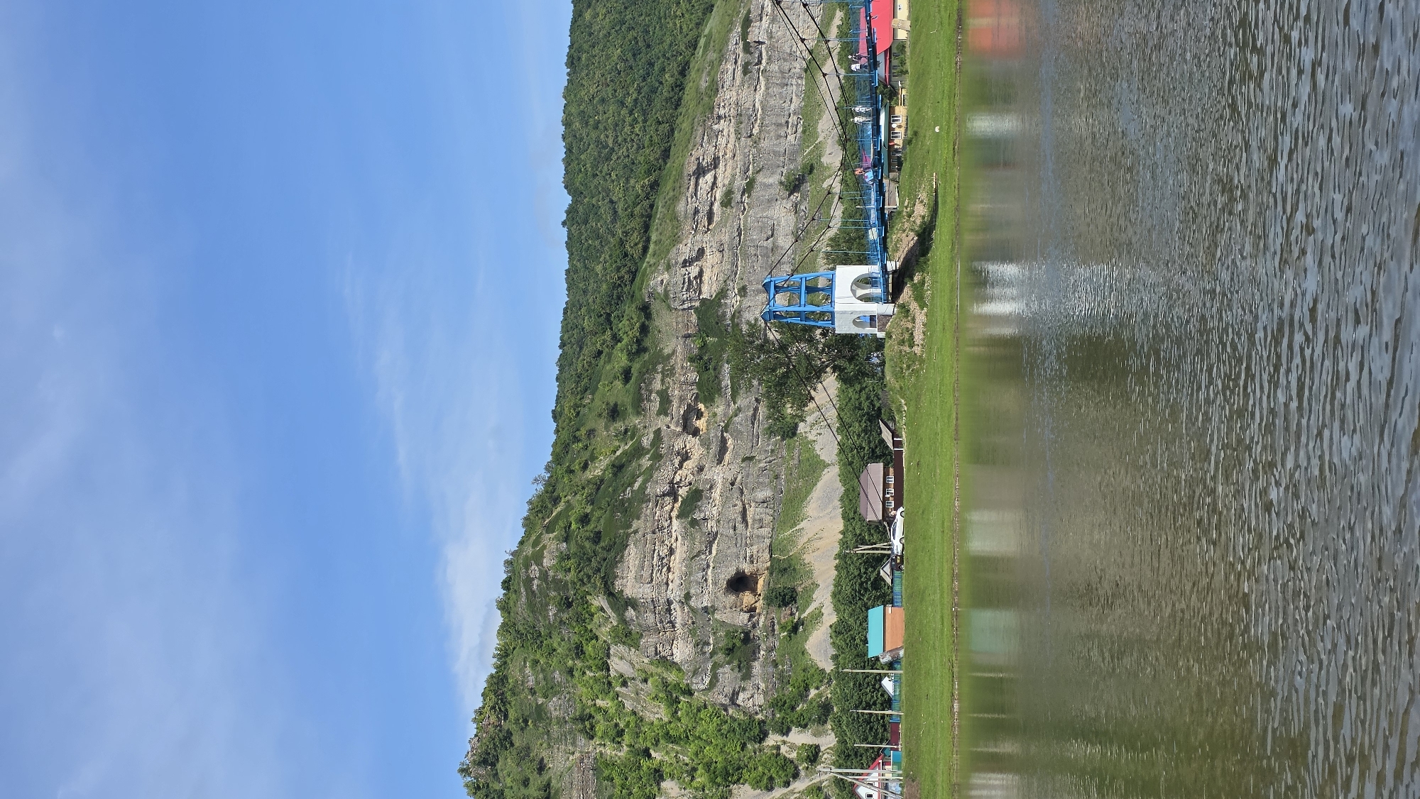 A panoramic, eye-level, full shot captures a scenic view of the Tashtyk River in Tash-Asty, Russia. The river occupies the foreground, its surface rippled and reflecting the sky and surrounding landscape. On the far bank, a lush green field slopes gently upwards, dotted with small, colorful houses featuring red and blue roofs. A white and blue suspension bridge with pedestrian traffic crosses the river, its towers a focal point. Behind the village, a massive, layered rock formation rises dramatically, its sheer face covered in dense green foliage. The sky above is a vibrant blue with scattered white clouds, indicative of a clear, sunny day.