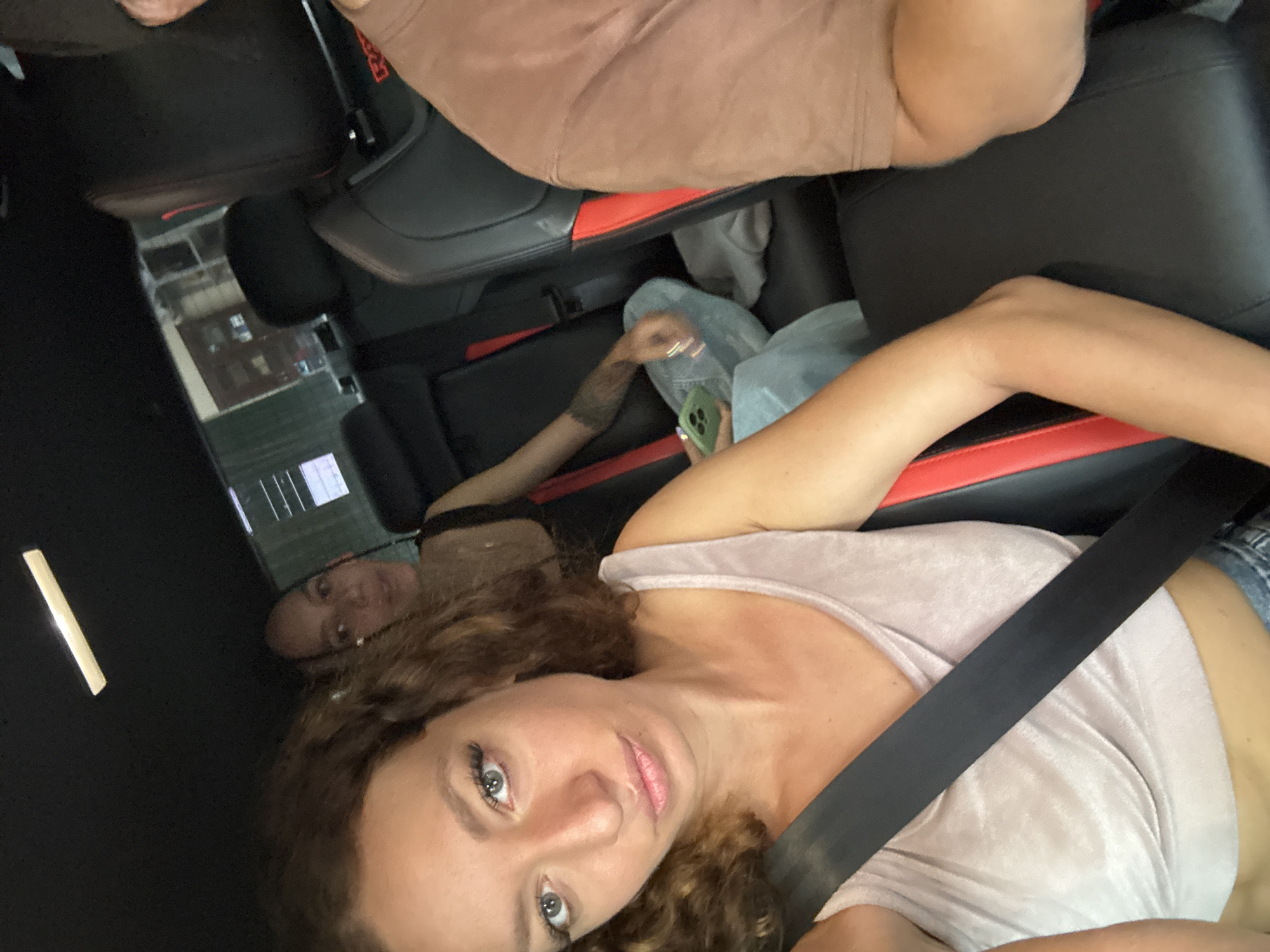Two people are in a car. One person, wearing a seatbelt and a light-colored top, is in the foreground, looking at the camera. The second person is behind them, holding a green phone. There are red accents on the car seats. Outside the car window, there is a view of buildings. The location appears to be in a city.