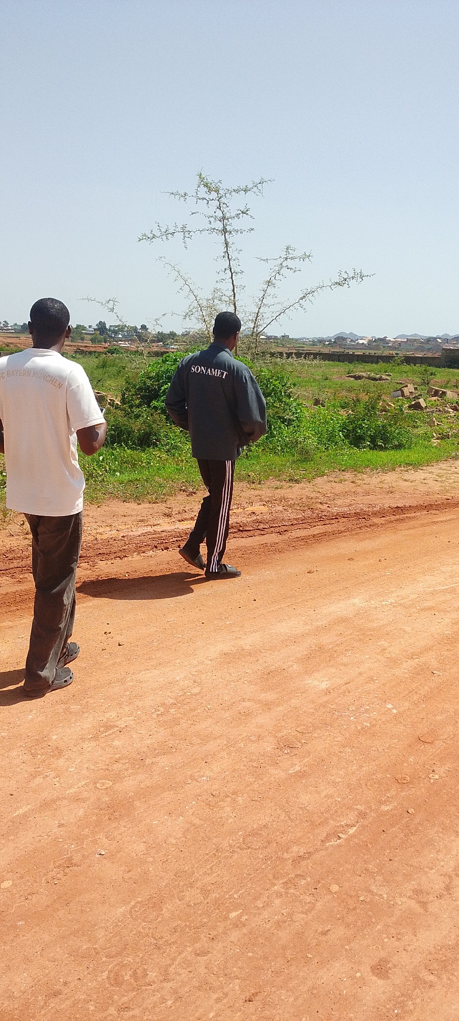 Two individuals are walking on a dirt road in Reifild-Ist, Nigeria. The person in the foreground on the left is wearing a white t-shirt with text that appears to say "FC BAYERN MUNCHEN." This individual is also wearing dark pants and dark shoes. The second person, walking ahead and to the right, is wearing a dark jacket with "SONAMET" printed on the back in white text. This person is also wearing dark pants with white stripes down the sides and dark shoes. The road is reddish-brown dirt, with tire tracks visible. To the right of the road is a grassy area with some bushes and scattered bricks. In the background, there are more trees, distant buildings, and a slightly hilly landscape under a clear sky. The overall environment suggests an undeveloped or semi-developed area.