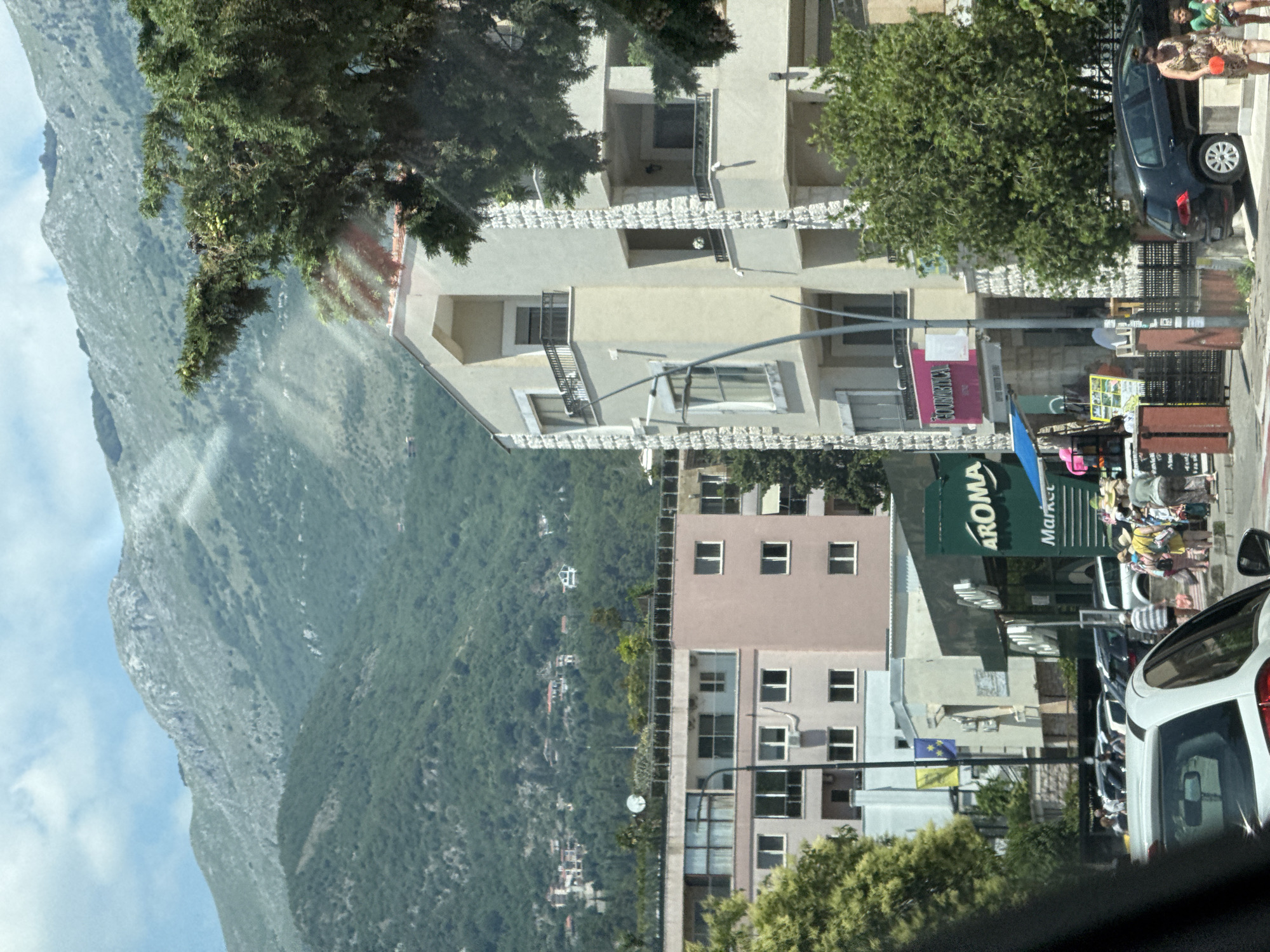 The image shows a street scene in Budva, Montenegro, with a view of mountains in the background. In the foreground, there are several buildings, including a light-colored apartment building with balconies and white stone accents. To the left, a pink apartment building is visible, and further to the left, a dark green tree. In the middle ground, there is a street with parked cars and pedestrians. A store called "AROMA Market" is visible, with a green facade. People are walking on the sidewalk and near the parked cars. The sky is partly cloudy.