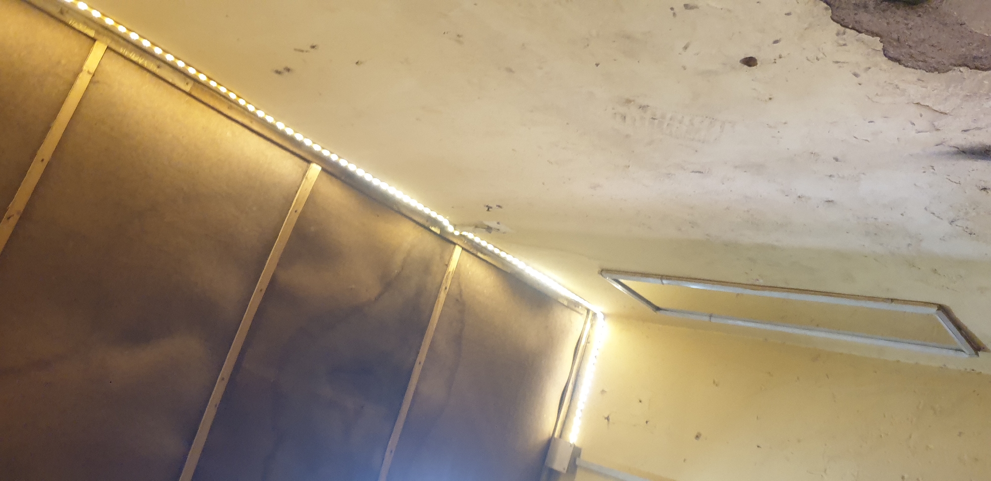 In Sabon Tasha, Nigeria, the image depicts the interior of a room with a textured, light-colored wall. A metallic frame, possibly for a window or door, is visible on the wall, oriented vertically and slightly angled. Along the ceiling, a strip of LED lights is installed, casting a warm, yellow glow downwards. The ceiling itself appears to be constructed of dark panels supported by metal framing. There are no people or animals visible in the frame. The overall impression is of an unfinished or utilitarian space with added lighting.