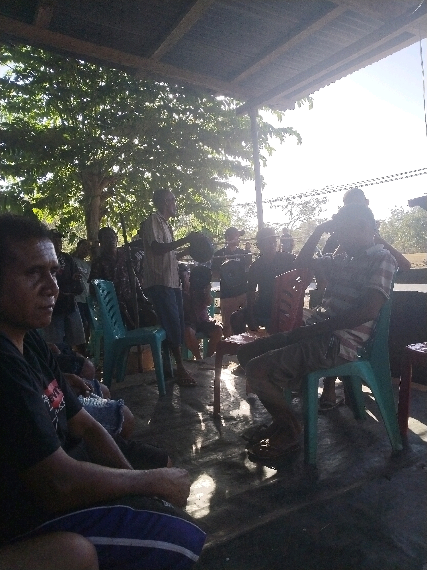 The image depicts a gathering of men under a covered outdoor structure. The setting appears to be Nggodimeda, Indonesia, given the context. Sunlight streams through trees, casting dappled shadows on the ground. Several men are visible, some standing and others seated on plastic chairs. One man in the foreground is looking to the left, his face partially obscured. Another man, standing, is holding a dark object, possibly a helmet, in his right hand. The men are casually dressed. The environment suggests a communal outdoor space, possibly a village area or a gathering place. The ground is uneven, and some debris is visible. The structure overhead seems to be made of wood and corrugated metal.