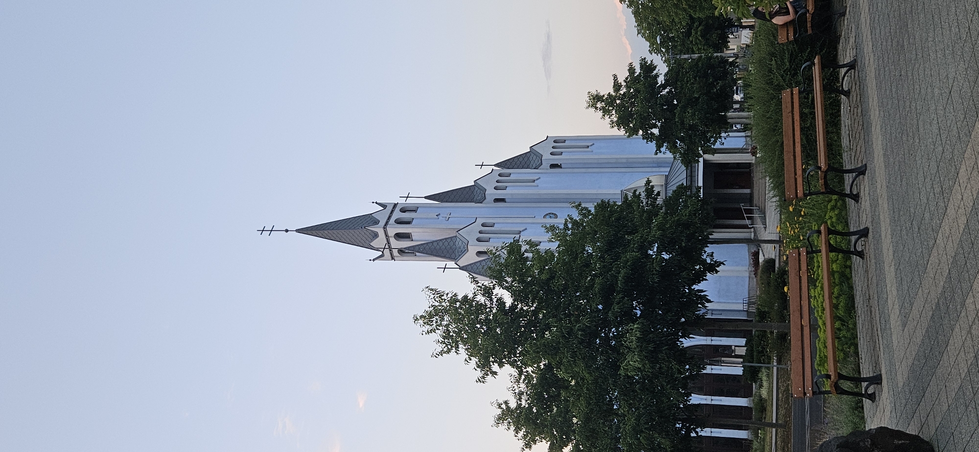The image shows a light blue church with a tall, pointed steeple and several smaller steeples. The church has a modern architectural style with sharp angles and a light blue facade. Trees with green foliage are in the foreground, and several wooden benches are visible on a paved area. A woman is seated on a bench on the right side of the image, looking at her phone. The sky is a pale blue with some wispy clouds. The church is identified as the Catholic Church of the Holy Spirit in Hévíz, Hungary.