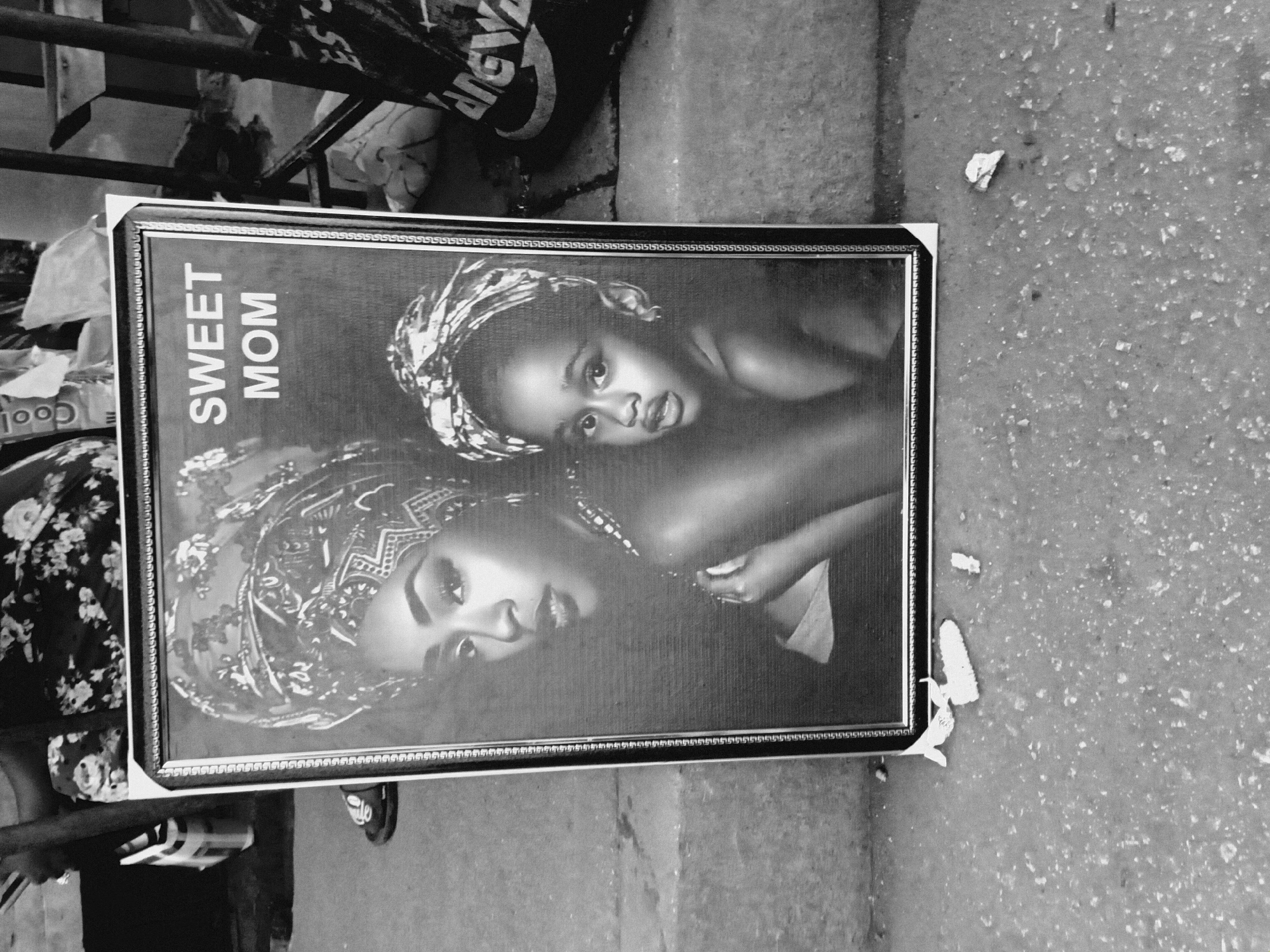 A black and white photograph depicts a framed portrait resting on a concrete surface outdoors. The portrait features an African woman and a child, both wearing head wraps. "Sweet Mom" is printed in sans-serif lettering on the left side of the portrait. The frame is dark with a decorative pattern. The background around the frame includes urban elements like metal bars, clothing, footwear, and scattered debris. The location is Bukuru, Nigeria.