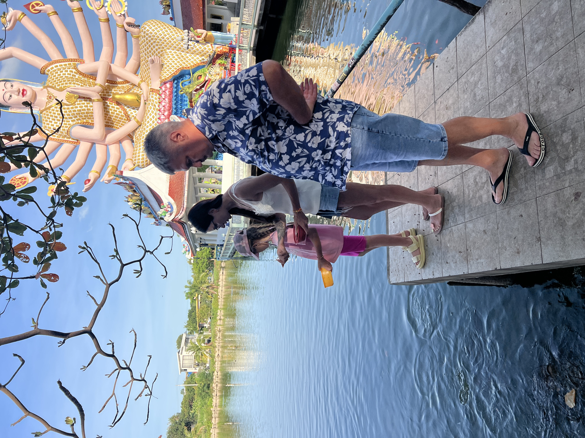 A family stands on a concrete platform overlooking a body of water in Baan Plai Laem, Thailand. The family consists of a man, a woman, and a young girl. The man wears a blue floral shirt and denim shorts, while the woman wears a white top and denim shorts. The girl wears a pink dress and a hat. They appear to be feeding fish in the water. A statue with multiple arms and a temple are visible in the background. The environment includes trees, buildings, and a clear blue sky.