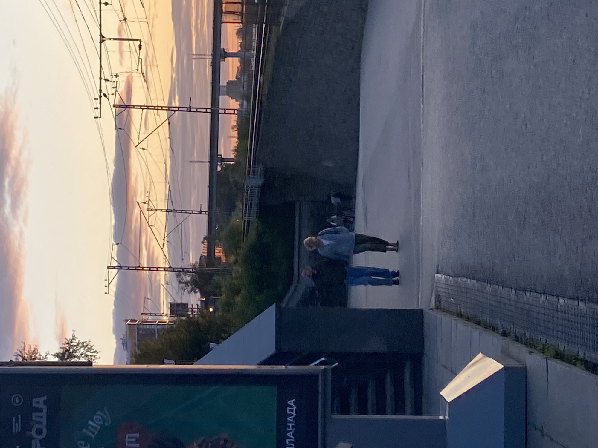 This image captures a street scene in Perm, Russia, during what appears to be sunset or dawn, given the warm hues in the sky. In the foreground, a man and a woman are walking side-by-side on a paved area. The man is wearing a dark jacket and blue jeans, while the woman is wearing a light blue jacket and dark pants. Behind them, a retaining wall made of large stones forms a boundary, topped by a railway track and overhead electrical lines, indicative of urban infrastructure.
To the left of the frame, a large, dark billboard or advertisement is partially visible. The text "РОДА" is clearly legible on the upper portion, suggesting a connection to the city's name or a local event. Further down, "ПЛАНАДА" is also visible. A dark, angular structure, possibly part of the billboard's mounting or an architectural element, stands in the foreground on the left, with a bright, reflective surface.
The background features a hazy skyline with buildings and trees, bathed in the soft light of the setting or rising sun, casting long shadows. The overall atmosphere is calm and transitional, with the city's elements gradually fading into the dusk.