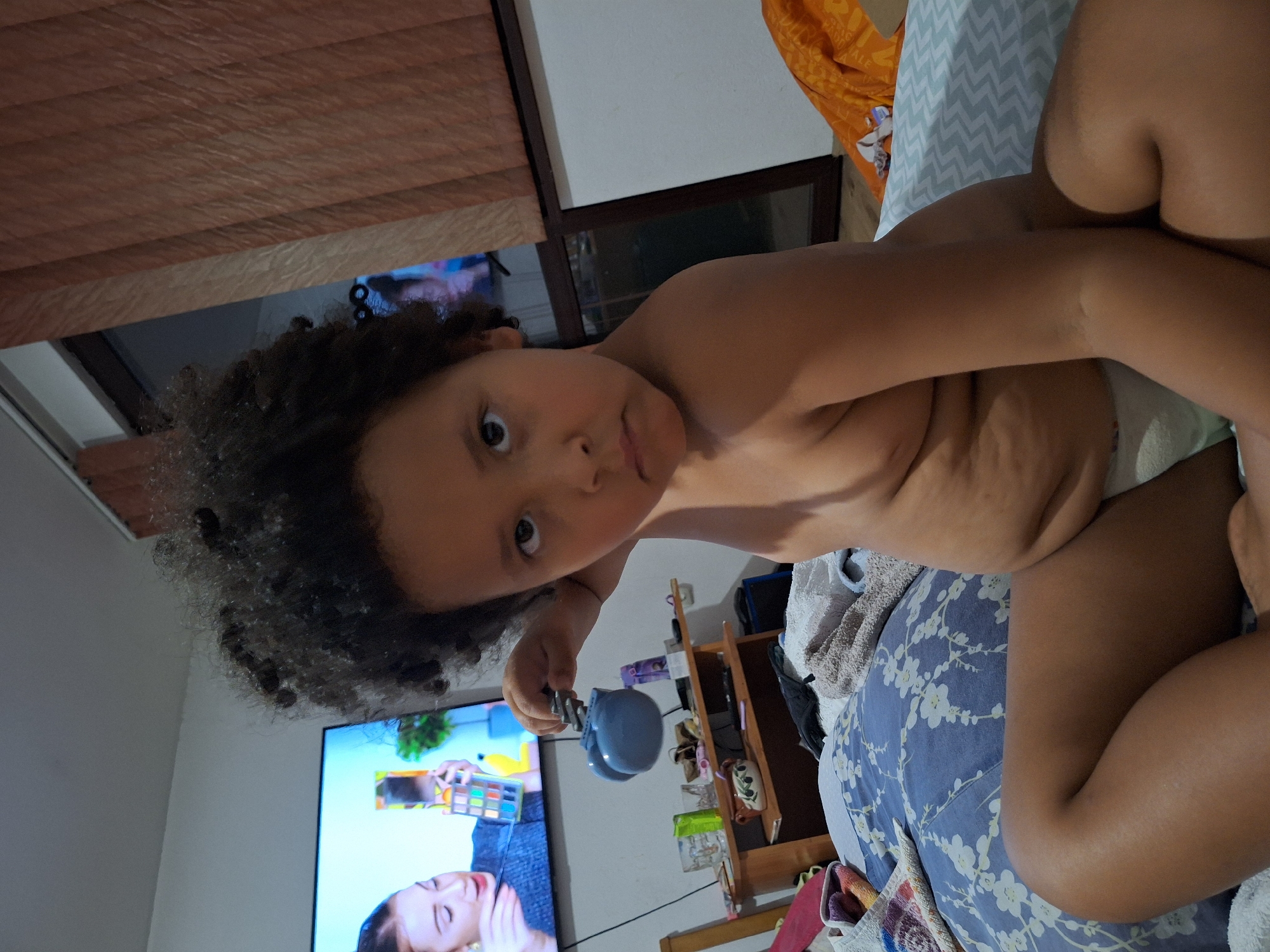 The image shows a young child lying on a bed. The child has curly, dark hair and is looking towards the camera with a neutral expression. The child is wearing a diaper and is shirtless. The background shows a television screen displaying a makeup tutorial, a shelf with various items, and a window with blinds. The room appears to be a bedroom. The overall lighting suggests it is daytime. The image was taken in Blagoevgrad, Bulgaria.
