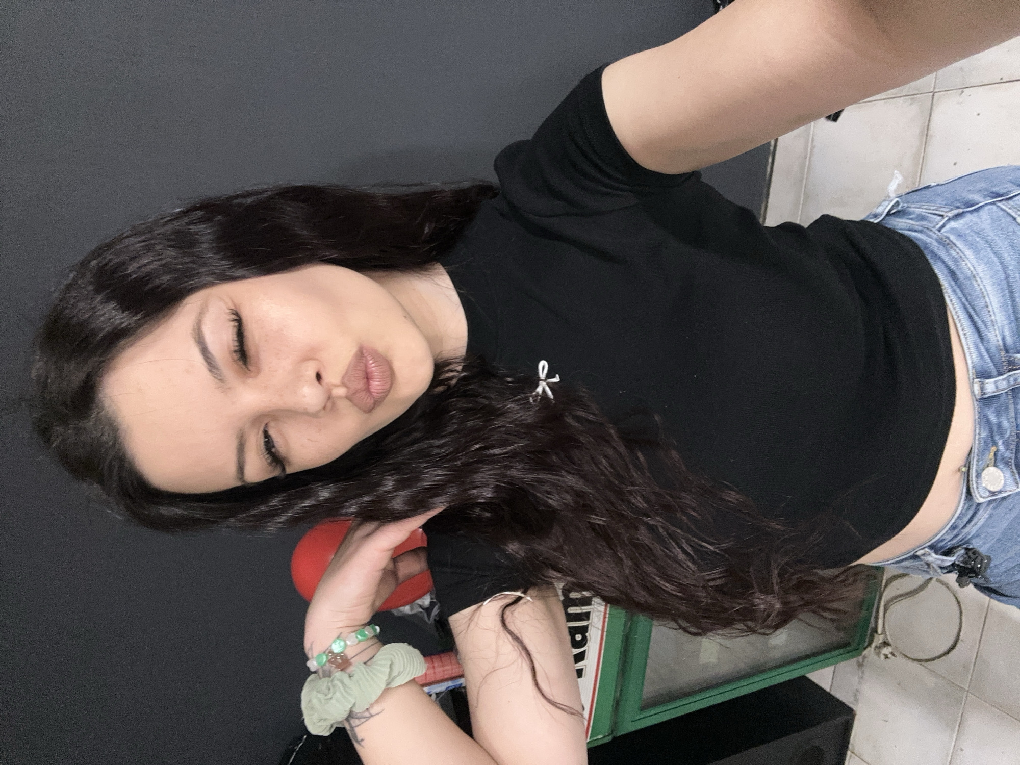 A woman with long, dark curly hair is posing for a selfie. She is wearing a black t-shirt and blue jeans. She has a bracelet and scrunchie on her left wrist. She is standing in front of a dark wall and a tiled floor. There is a red object and a green metal frame with a glass insert in the background. There is no information available to determine if the photo was taken in Sliven, Bulgaria.