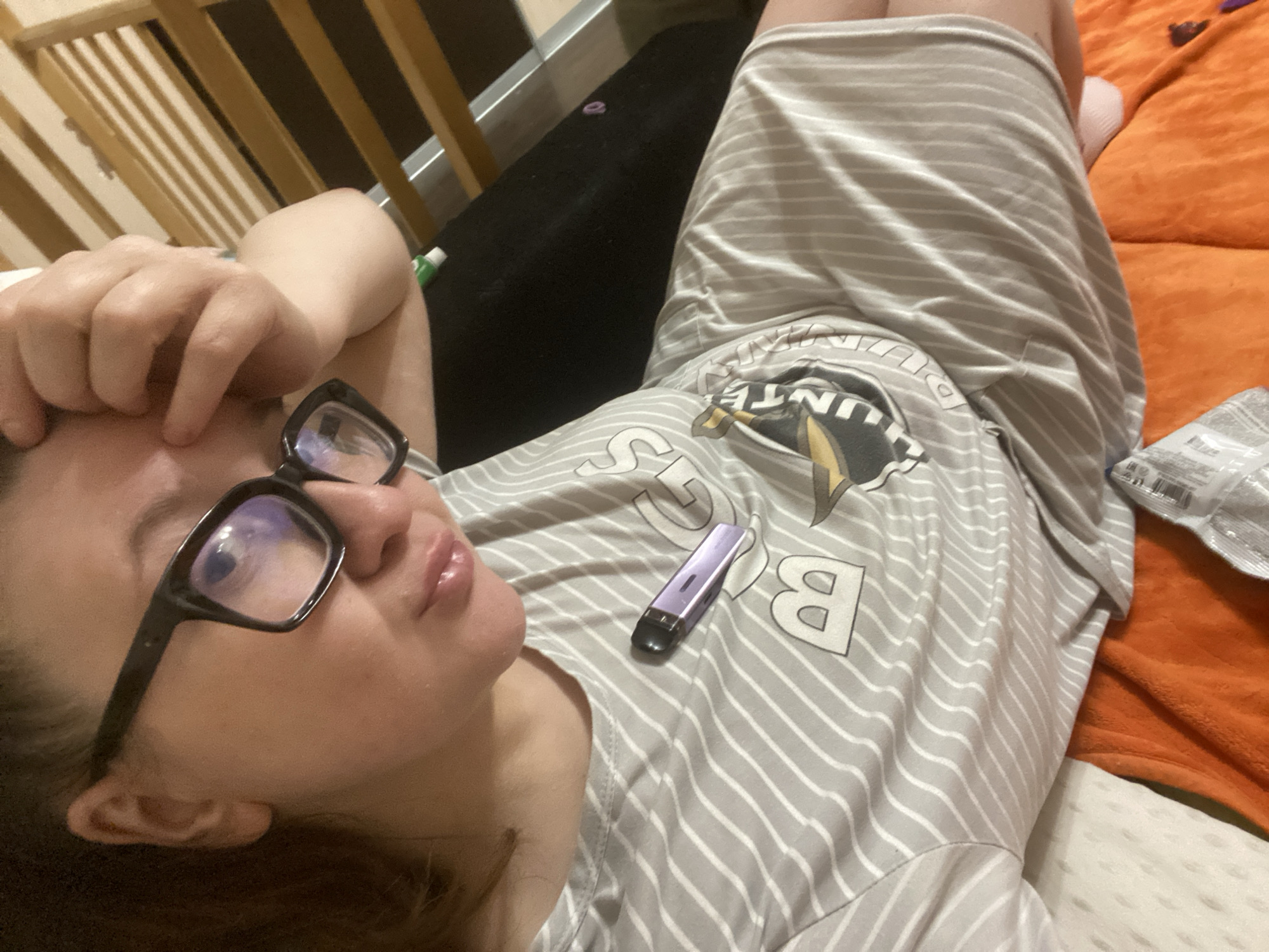 A person is lying down, wearing glasses and a grey and white striped shirt. There is a purple vape device resting on the shirt. An orange blanket is visible on the right side of the frame. A wooden structure is in the background to the left. The location and city are not discernible from the image.