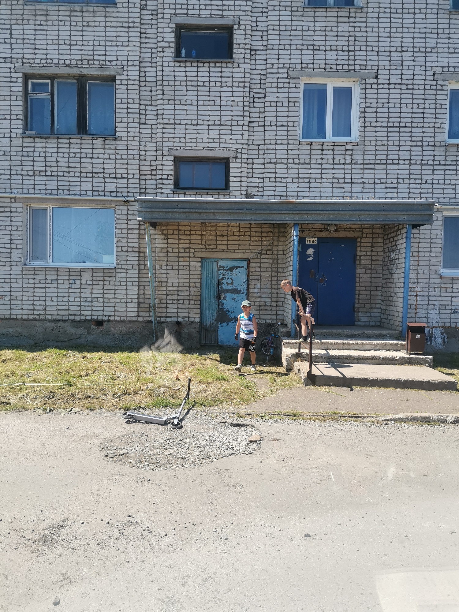 In Vanino, Russia, a low-angle, eye-level shot captures the facade of a multi-story brick building. The building exhibits a utilitarian design with numerous rectangular windows, some white-framed and others dark. A covered entryway with a corrugated metal roof is supported by blue metal poles. A blue door, appearing weathered, is visible beneath the awning. To the left of the entrance, a smaller, equally distressed blue door is present.
In front of the building, two young boys are present. One boy, wearing a striped tank top and dark shorts, stands on the grass to the left, facing towards the entrance. Beside him, a small bicycle leans against a railing. The second boy, dressed in a black t-shirt and dark shorts, stands on the steps leading up to the blue door, his body angled downwards as if looking at something on the ground or at the other boy.
In the foreground, a grey scooter lies on the asphalt, angled towards the right of the frame. The ground in front of the building is a mix of cracked asphalt and patches of gravel, with a patch of dry grass and weeds between the road and the building's foundation. A small, brown metal trash receptacle is positioned on the right side of the steps leading to the entrance. The scene suggests a casual outdoor setting on a bright, sunny day.
