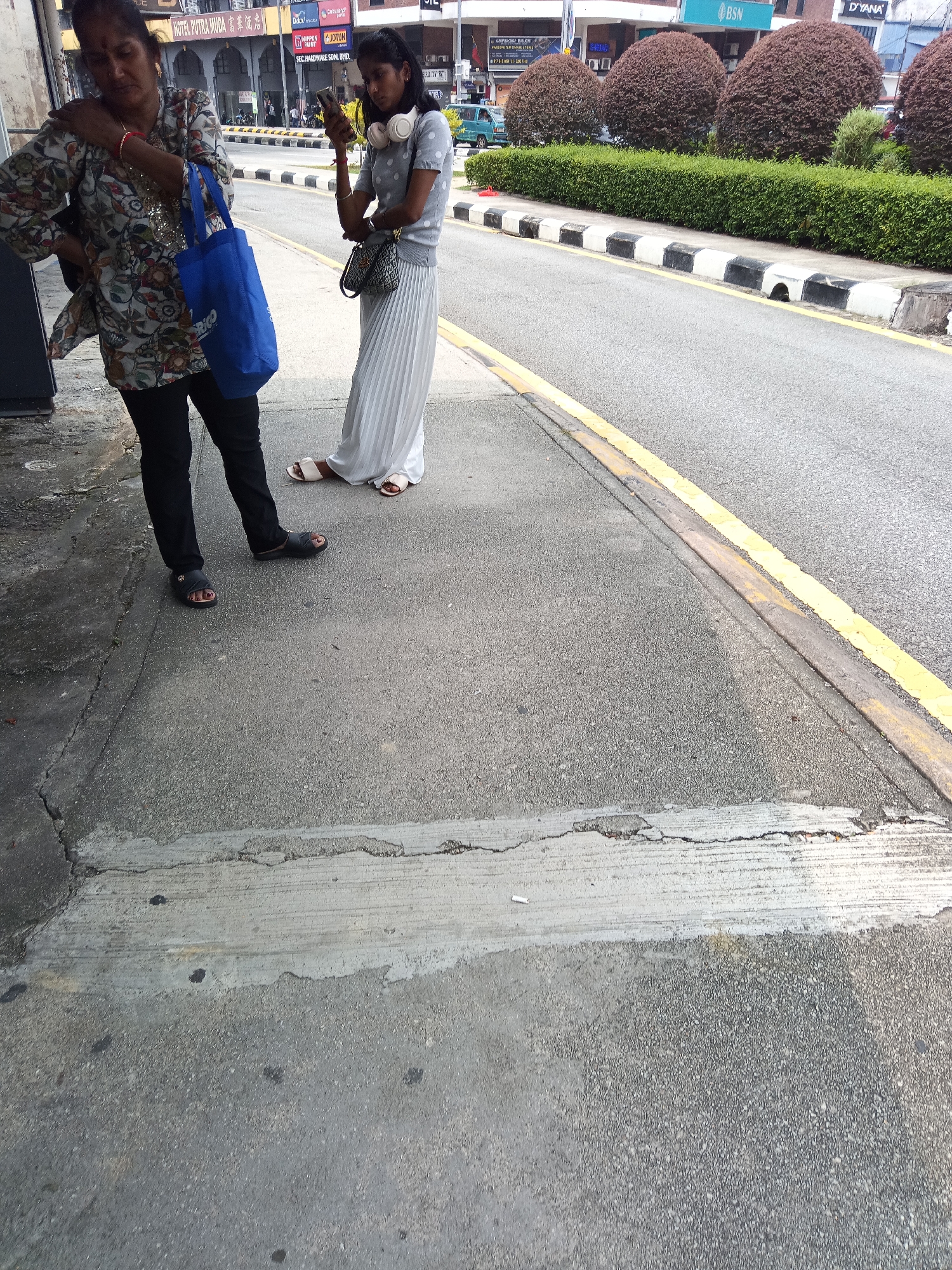 Two women are standing on a sidewalk next to a street in Kuala Lumpur, Malaysia. The woman on the left is wearing a patterned shirt and black pants, and is holding a blue shopping bag. She has her right hand on her shoulder and her left hand by her side. The woman on the right is wearing a light-colored polka dot shirt and a long, pleated white skirt. She is holding a mobile phone in her right hand and wearing headphones around her neck. She is looking down at her phone. The sidewalk is made of concrete and is cracked in places. The street is paved with asphalt and has a yellow line painted along the edge. In the background, there are buildings with signs, including one that says "BSN" and another that says "D'YANA." There are also manicured bushes and trees along the side of the road. The scene appears to be outdoors during daylight hours.