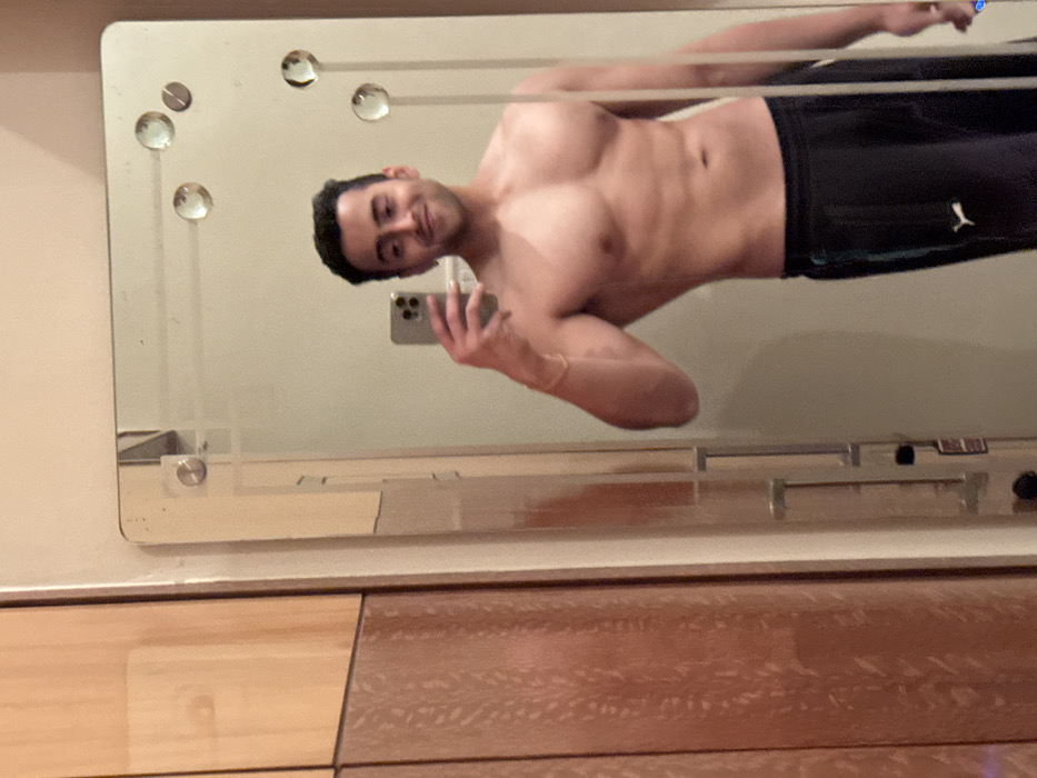 A shirtless man in black athletic shorts with a white Puma logo is seen in a mirror selfie. He is holding a phone in his right hand, angled toward himself, with a partial view of his face and torso. The mirror has decorative glass elements at the top and is mounted above a wooden surface, possibly a dresser or cabinet. The man has dark hair and a light complexion.