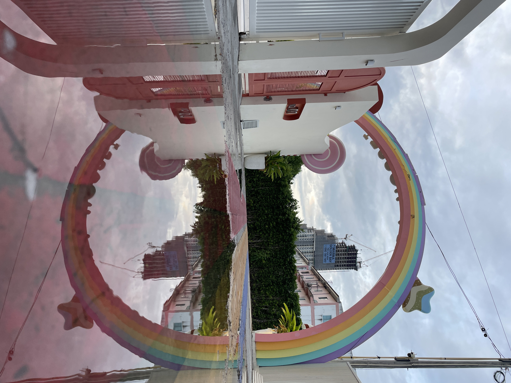 A low-angle outdoor shot shows a reflection in a puddle of a whimsical archway in Pattaya City, Thailand. The arch is shaped like a rainbow with a star-shaped decoration on top. To the left of the arch, a white building with a red door and the house number "01" is visible. Next to the building is a large pink lollipop-shaped decoration. A lush green hedge forms a backdrop behind the structures. In the distance, a tall building under construction is partially visible, with a banner advertising "MARINA GOLDEN BAY". The sky is overcast with clouds. The puddle reflects the archway, the building, the hedge, and the sky, creating a mirrored image of the scene.