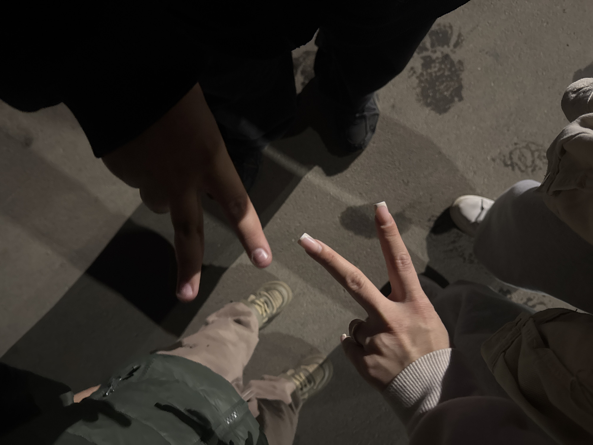 The image shows an aerial view of three people standing on a concrete surface. Two of the individuals are displaying the "peace" or "victory" hand gesture. One person is wearing dark clothing and black shoes. Another is wearing tan pants and light-colored shoes. The third individual is wearing beige pants and white sneakers. The location cannot be determined without further information. The presence of concrete suggests an urban environment.