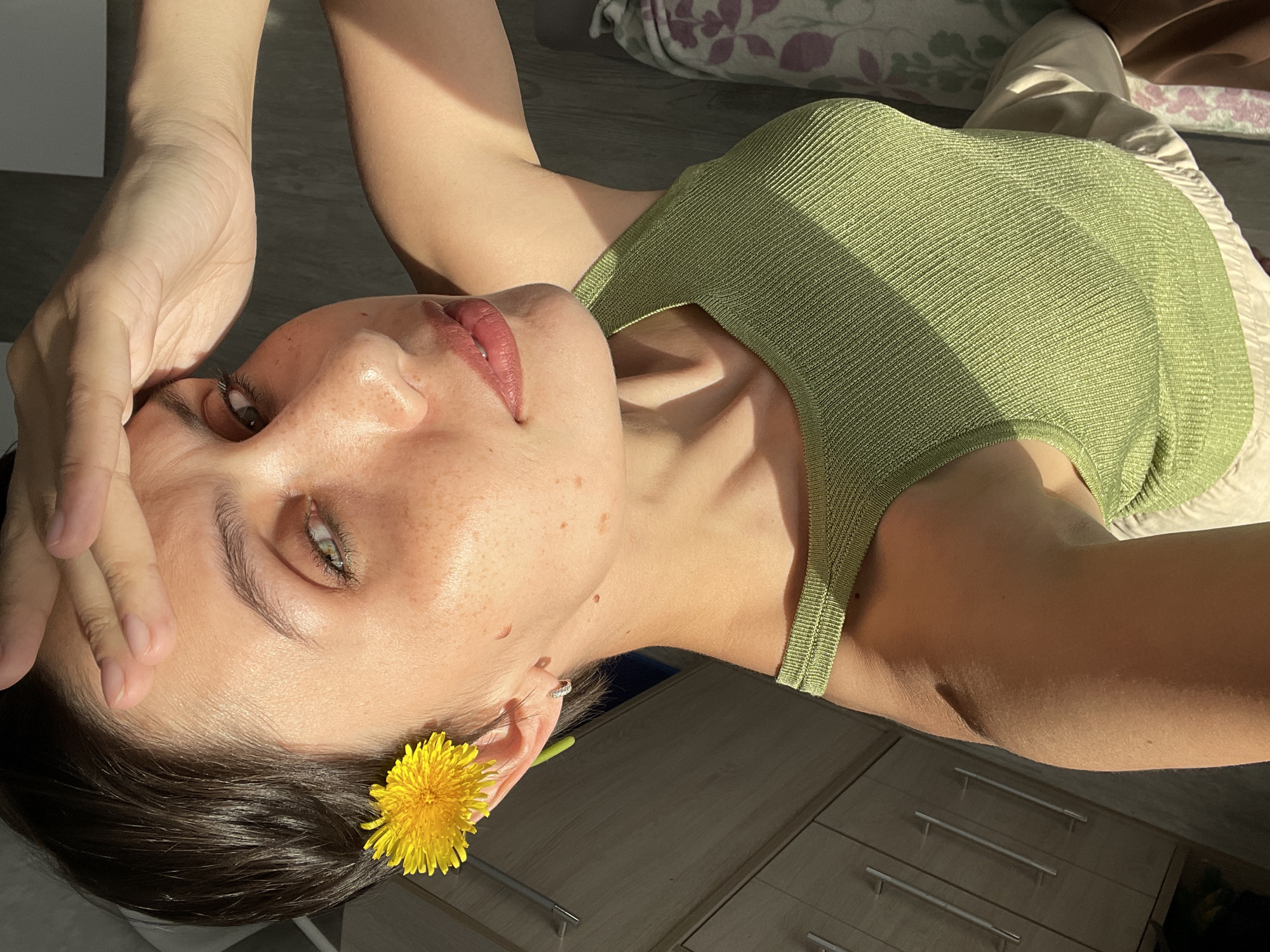 A young woman with light skin, freckles, dark hair, and green eyes poses for a selfie. She is wearing a light green ribbed tank top and light-colored pants. A yellow dandelion is tucked behind her left ear. Her left hand is raised near her forehead. She is lying on a bed with floral patterned bedding and appears to be in a room with light-colored wooden floors and furniture, which appear to be drawers. No evidence suggests the image was taken in Izhevsk, Russia.