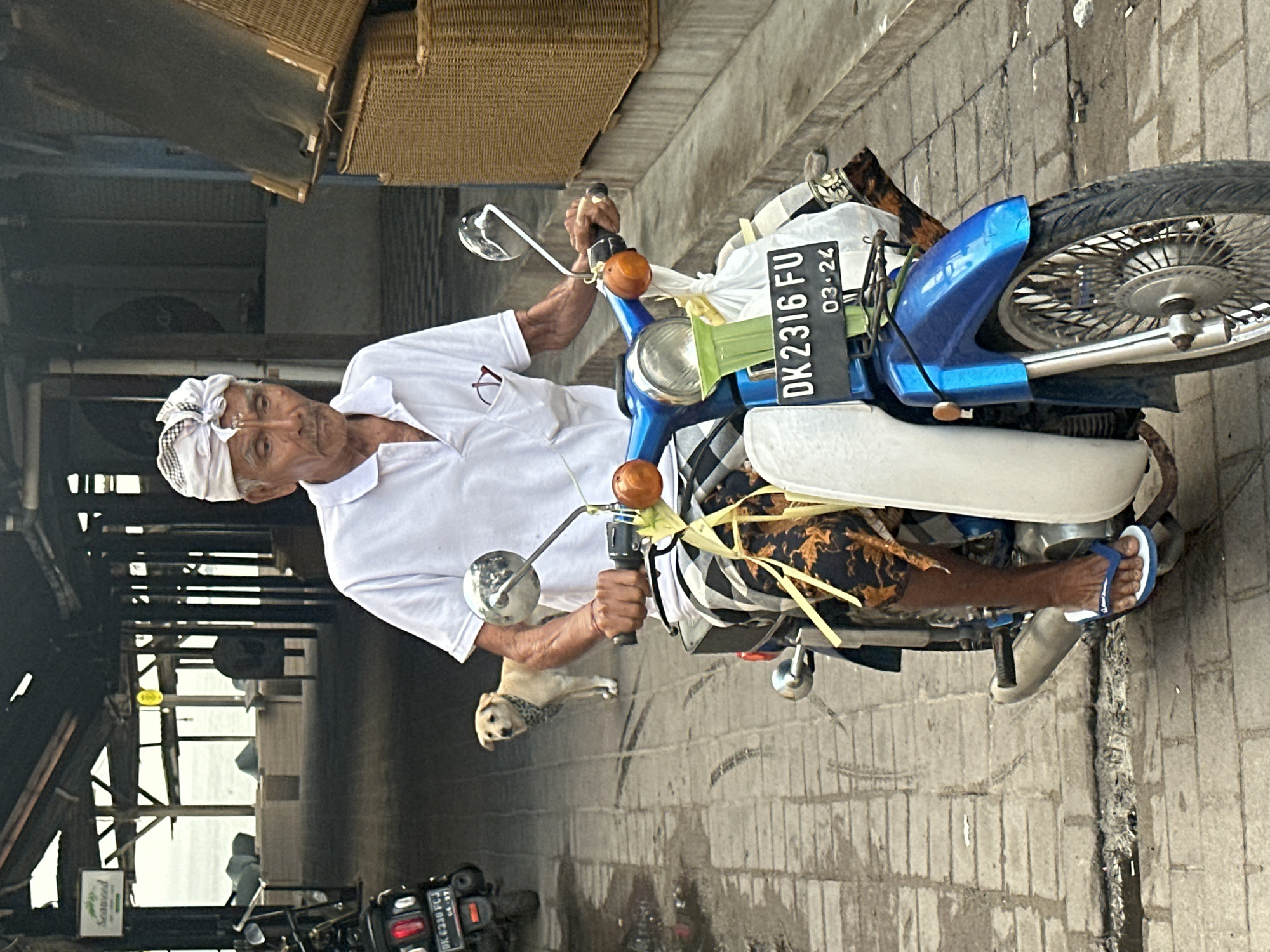 An elderly Indonesian man, wearing a white polo shirt and a white bandana, sits on a blue and white motorcycle. He has a thin build and is wearing flip-flops. The motorcycle has a license plate that reads "DK 2316 FU 03-24". In the background, a light-colored dog stands on a paved street. The scene appears to be in an outdoor market or a street in North Kuta, Indonesia, with buildings and structures visible behind the man and dog. There are woven baskets on the right side of the image.