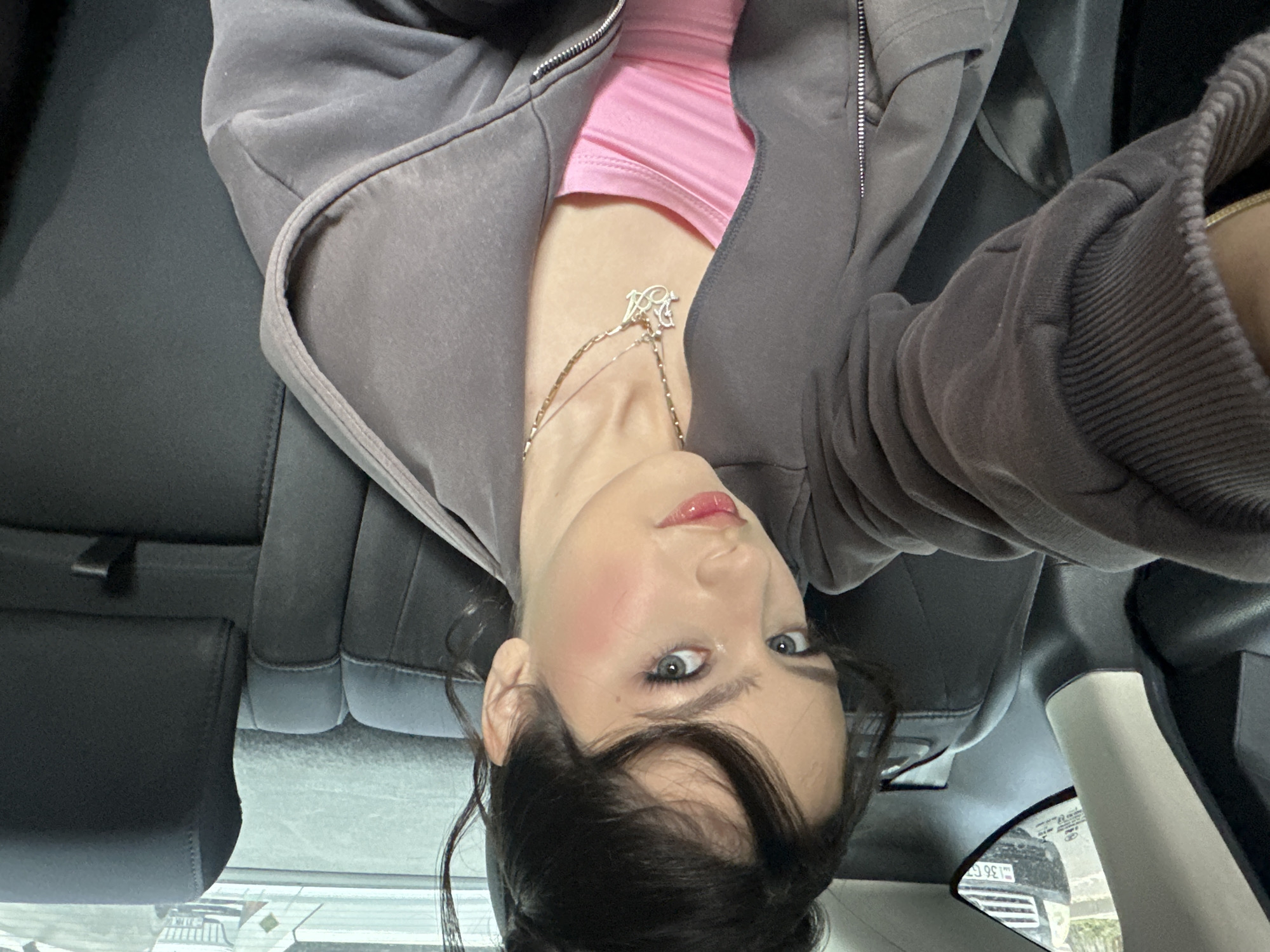 A young woman is in the back seat of a car. She is wearing a pink top under a gray zippered hoodie, and a silver necklace. The car is parked, as evidenced by the view of the street outside the window. The license plate visible in the reflection of the car parked behind them seems to indicate the location is in Yerevan, Armenia.