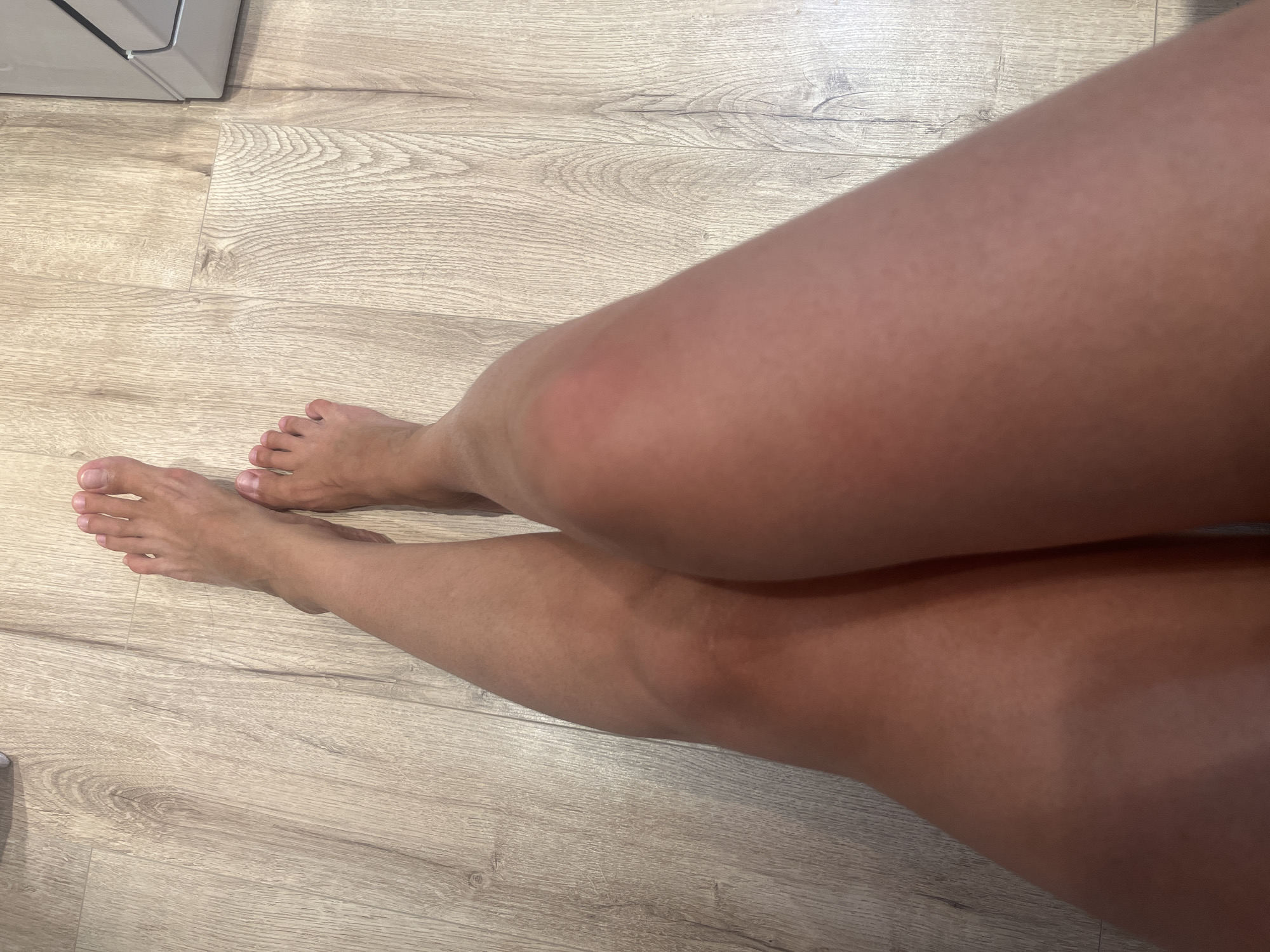 The image shows a close-up, overhead view of a person's bare legs and feet resting on a light-colored wooden floor. The person's skin appears tanned. The left leg is positioned across the right leg. The toes of both feet are visible, with toenails appearing unpolished. The floor is made of wood planks with a visible grain pattern. Part of a white appliance, possibly a washing machine or refrigerator, is visible in the upper right corner of the image. The scene is indoors, and there is no indication of the specific location within the city of Rybnitsa, Moldova.