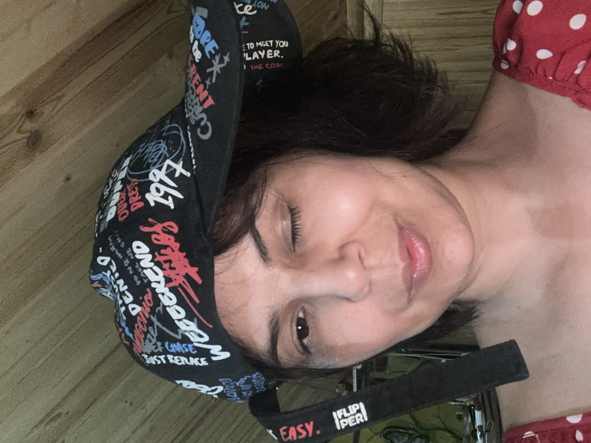 A woman is shown in a close-up, medium shot, wearing a black baseball cap with a graffiti-style print. She is looking at the camera and winking with her right eye. The cap has various words and phrases printed on it, including "WEEKEND," "DENIED," "1997," and "EASY." There is also a black strap hanging from the cap, with the words "FLIP PER" printed on it. The woman has dark, chin-length hair and is wearing a red top with white polka dots. The background appears to be a wooden wall. The image does not provide enough information to determine the location in the city of Kalininskaya, Russia.